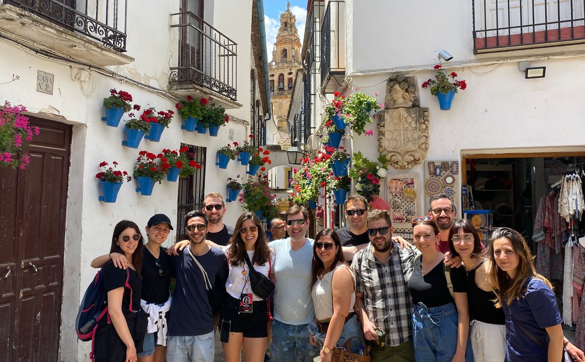 A WeRoad group trip poses for a photo on a narrow street with white walls adorned with blue flower pots and a bell tower in the background.