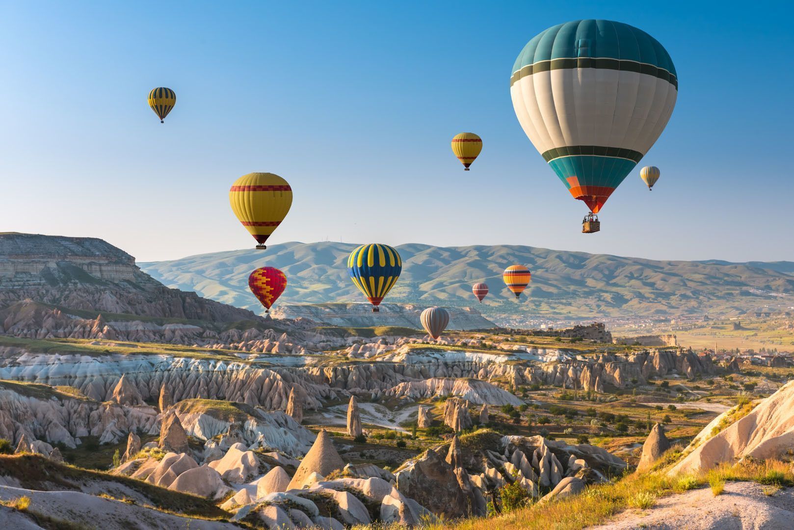 Globos aerostáticos de colores vuelan sobre un vasto paisaje rocoso con formaciones cónicas bajo un cielo despejado.