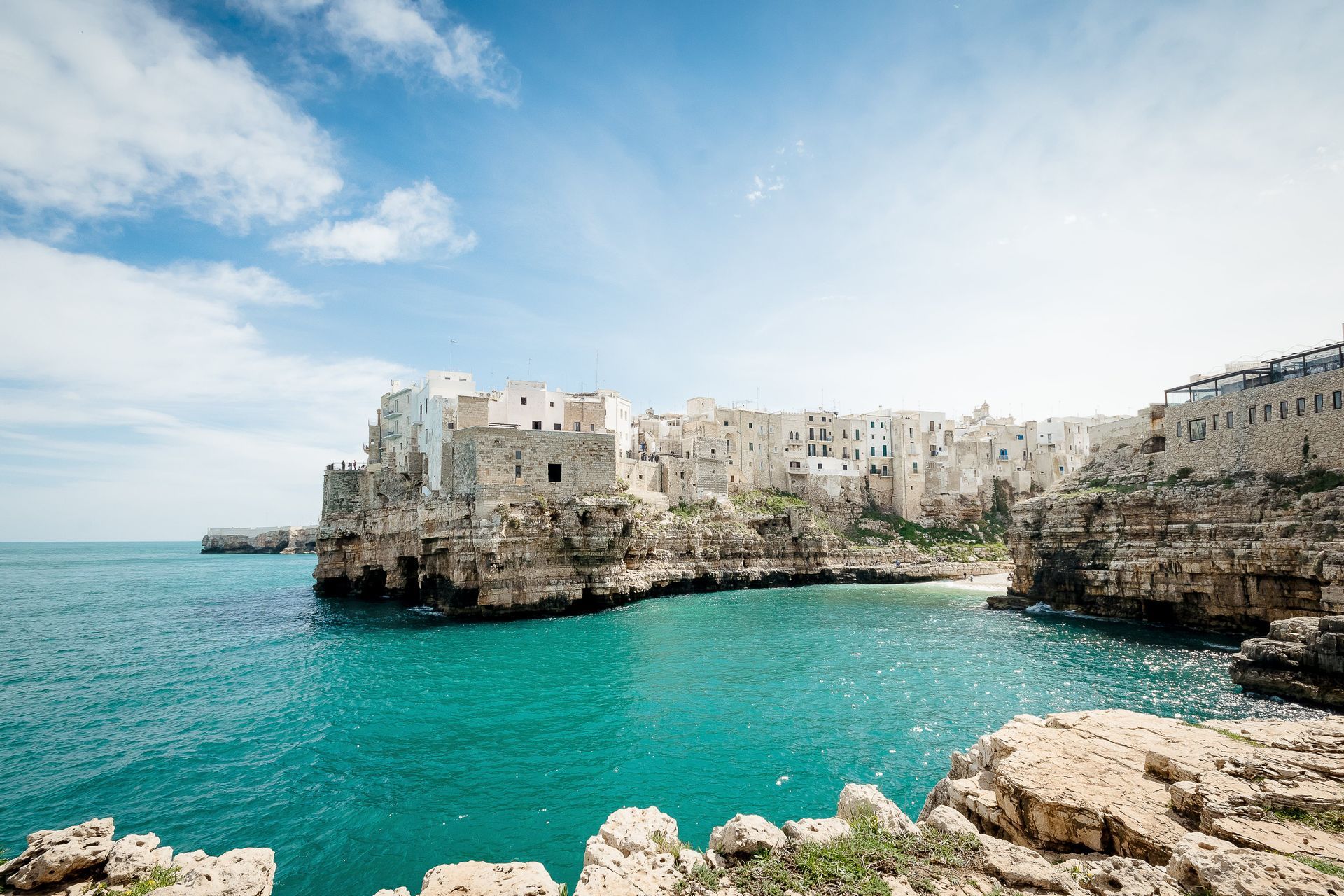 Une ville aux bâtiments blancs et en pierre est perchée au sommet d'une falaise rocheuse surplombant une baie turquoise sous un ciel bleu parsemé de nuages.