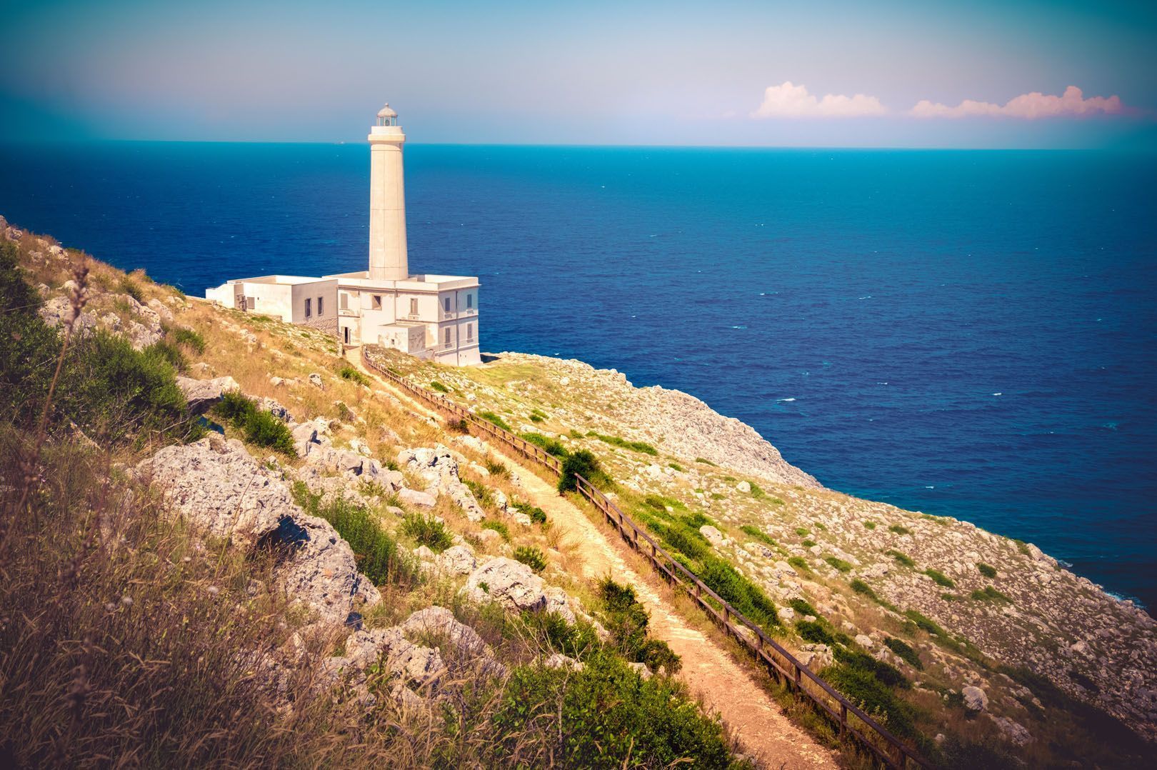 Un faro blanco y alto sobre un acantilado cubierto de hierba, con vistas al mar azul profundo y un sendero de tierra en primer plano.