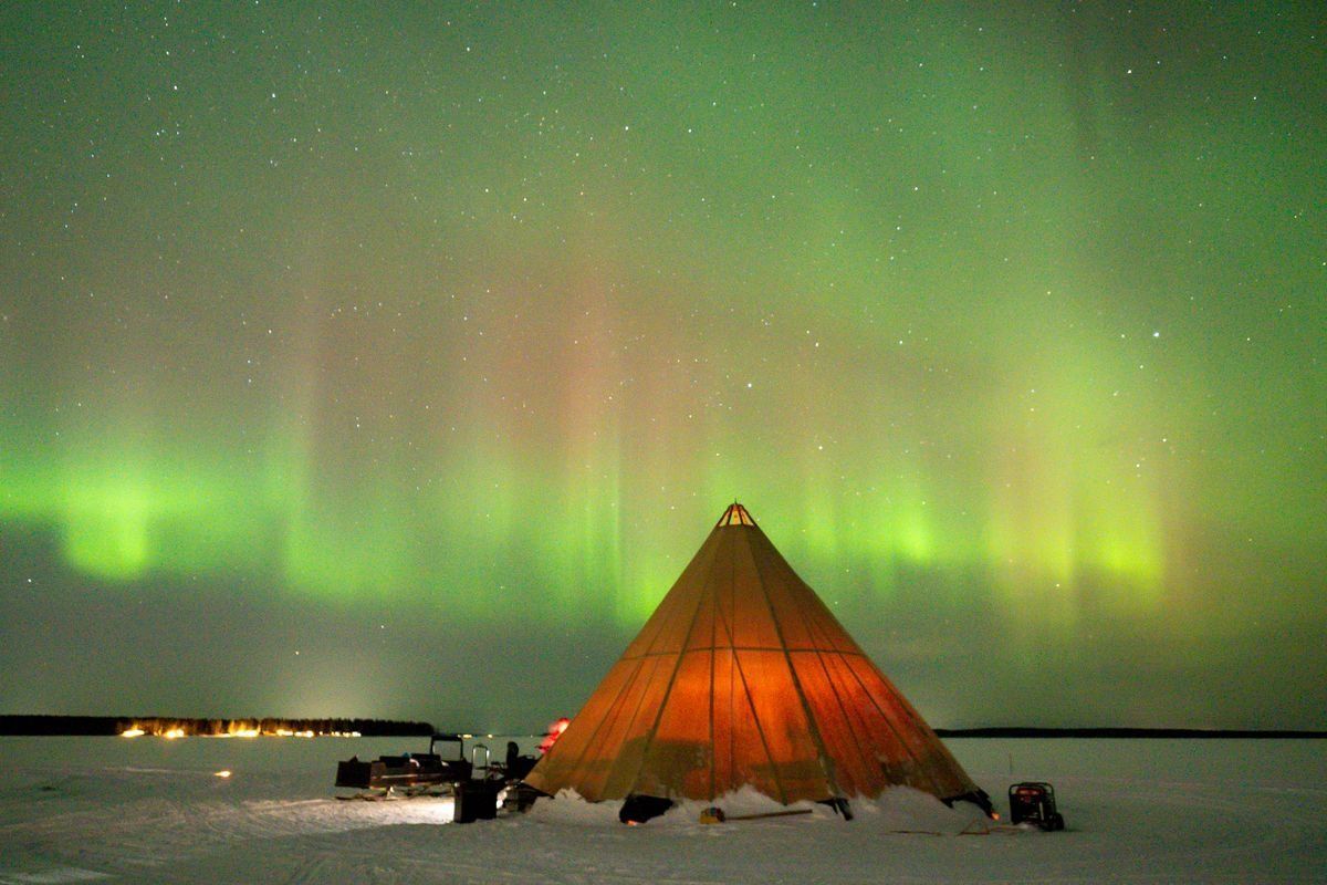 Un tipi illuminato si erge in un paesaggio innevato sotto un cielo notturno stellato, avvolto dalla verde Aurora Boreale.