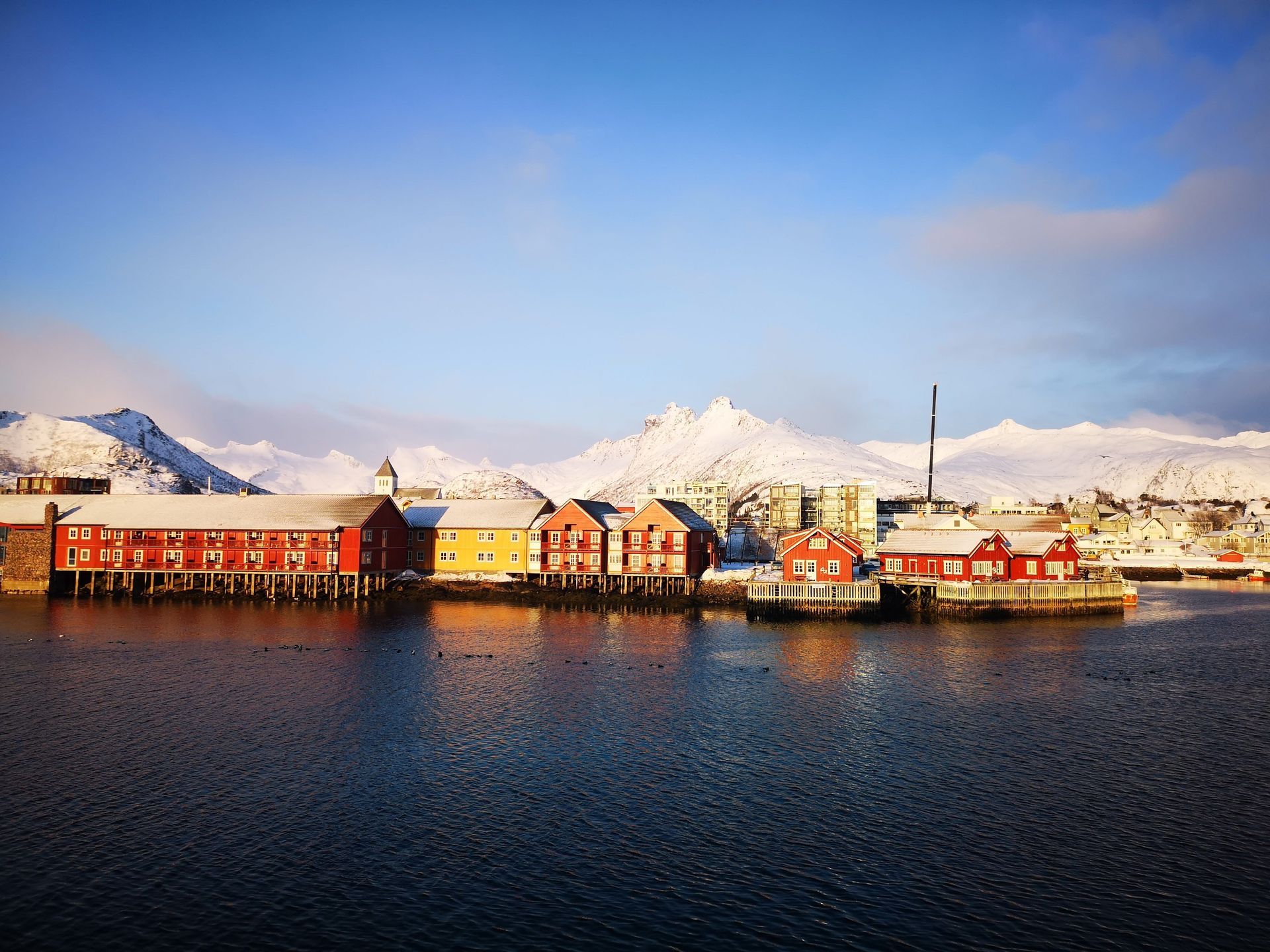 Des maisons colorées rouges et jaunes sur pilotis bordent un port calme avec de grandes montagnes enneigées en arrière-plan sous un ciel bleu.