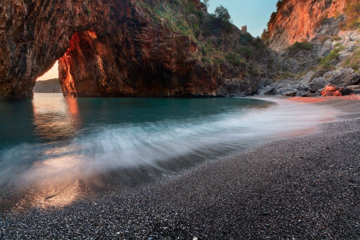 Una foto a lunga esposizione di un'onda che si infrange su una spiaggia di ciottoli scuri in una caletta con un grande arco roccioso illuminato dalla luce del tramonto.