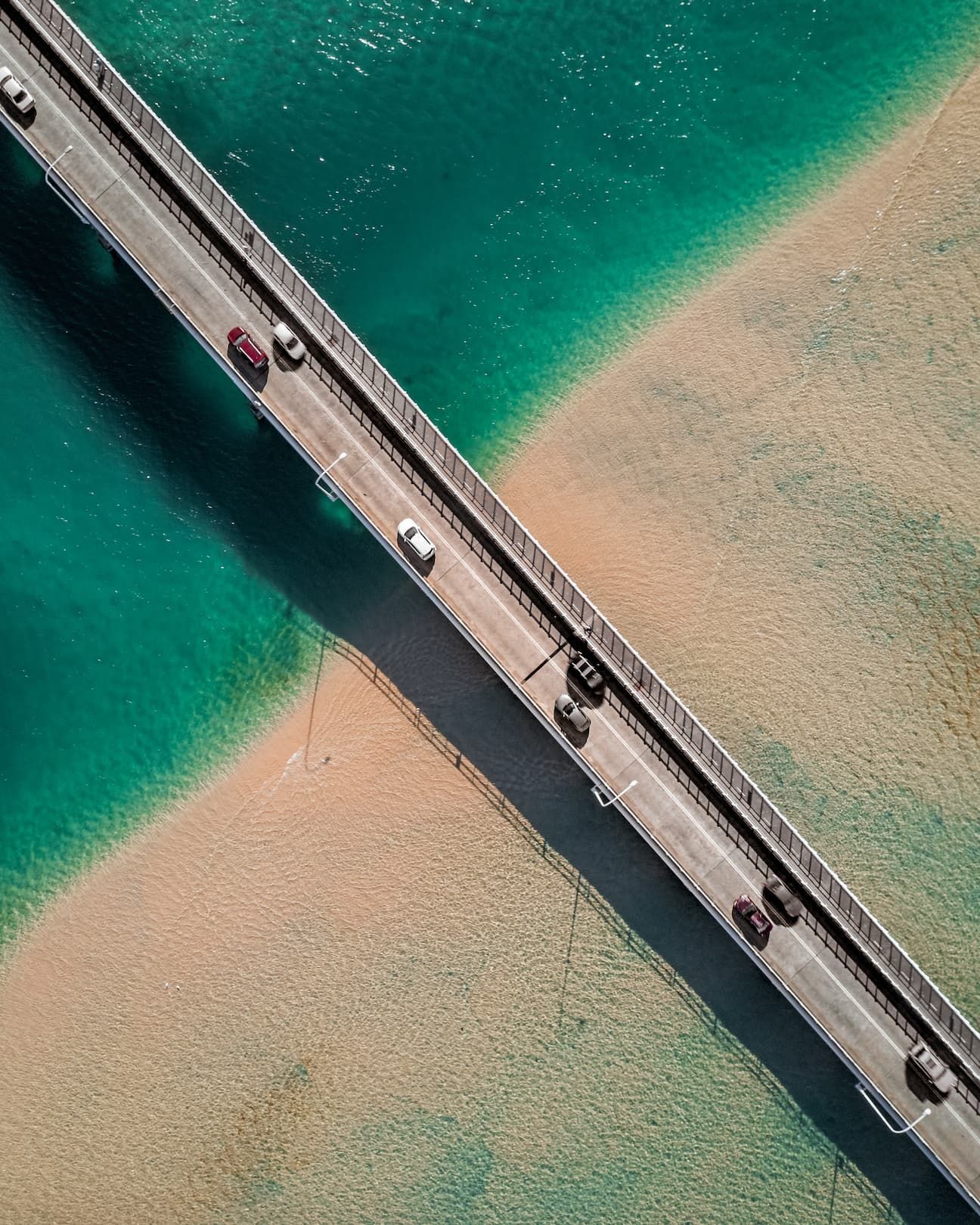 An aerial top-down view of cars driving on a bridge crossing over turquoise water and a sandy shoreline.
