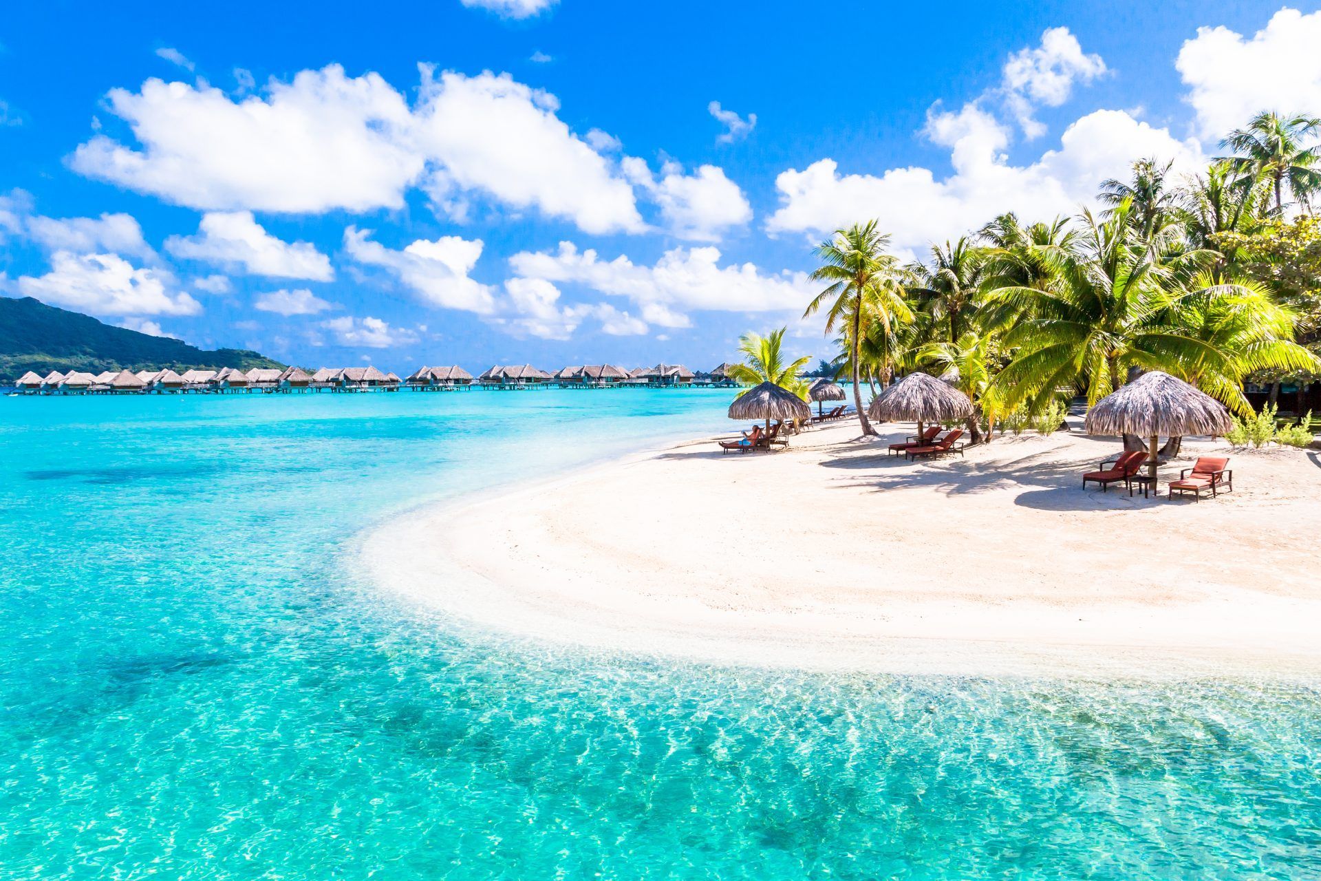 Bungalow sull'acqua su acque turchesi, accanto a una spiaggia di sabbia bianca con palme e ombrelloni sotto un cielo blu.