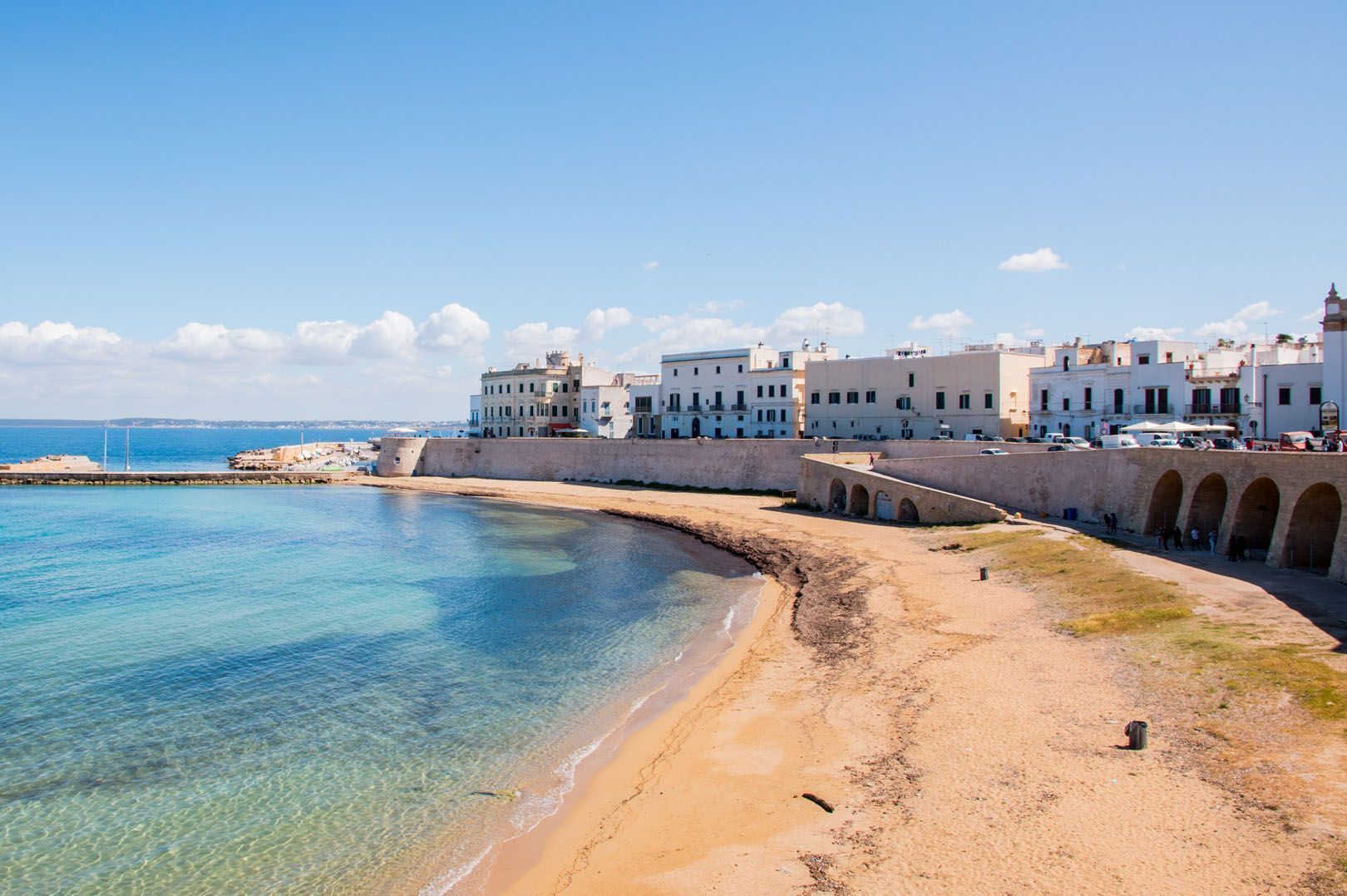 Una spiaggia di sabbia con acque cristalline turchesi si estende lungo una storica città costiera, caratterizzata da edifici bianchi e un muro marittimo in pietra.