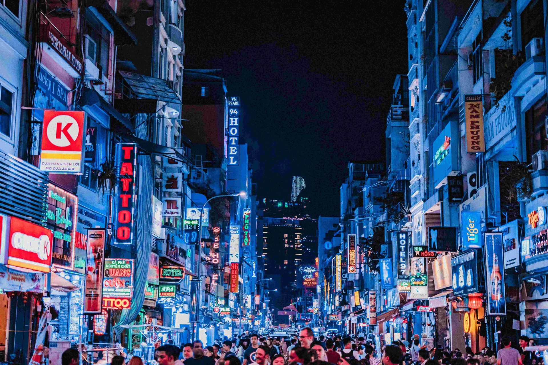 Eine belebte Stadtstraße bei Nacht, auf der Menschen unter hohen Gebäuden mit leuchtenden blauen und roten Neonschildern entlanglaufen.