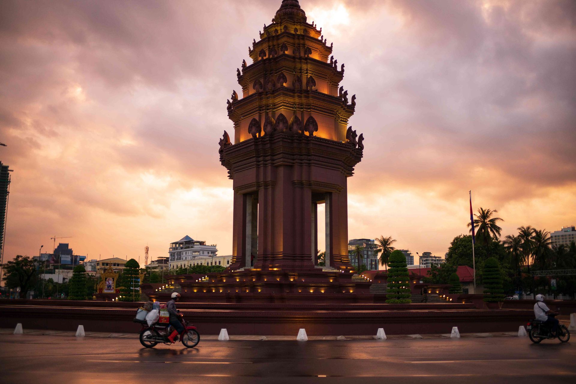 Un monument orné et illuminé se dresse sur une place de ville au coucher du soleil, tandis que deux motos passent sur une route mouillée.