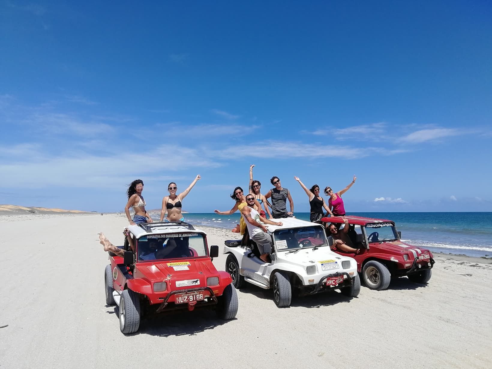 Un viaggio di gruppo WeRoad in posa su tre veicoli fuoristrada su una spiaggia sabbiosa con l'oceano e un cielo blu sullo sfondo.