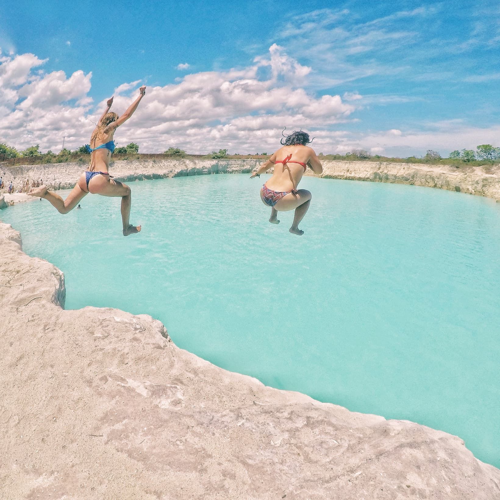 Dos mujeres en bikini saltando desde un saliente rocoso a una piscina de agua turquesa bajo un cielo azul y nublado.