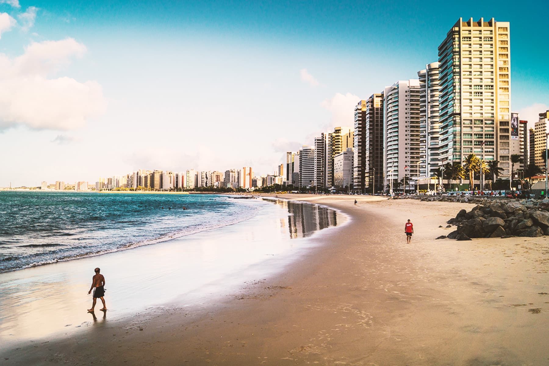 Una vasta spiaggia sabbiosa con poche persone che passeggiano lungo la riva, delimitata da un moderno skyline urbano con numerosi grattacieli.