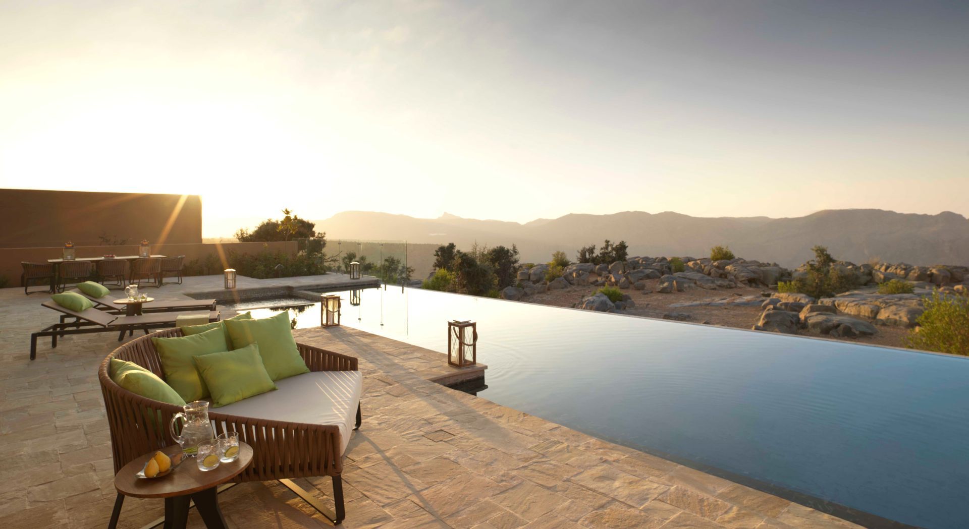 Un patio moderno con piscina a sfioro e mobili lounge che si affaccia su un paesaggio montano roccioso al tramonto.