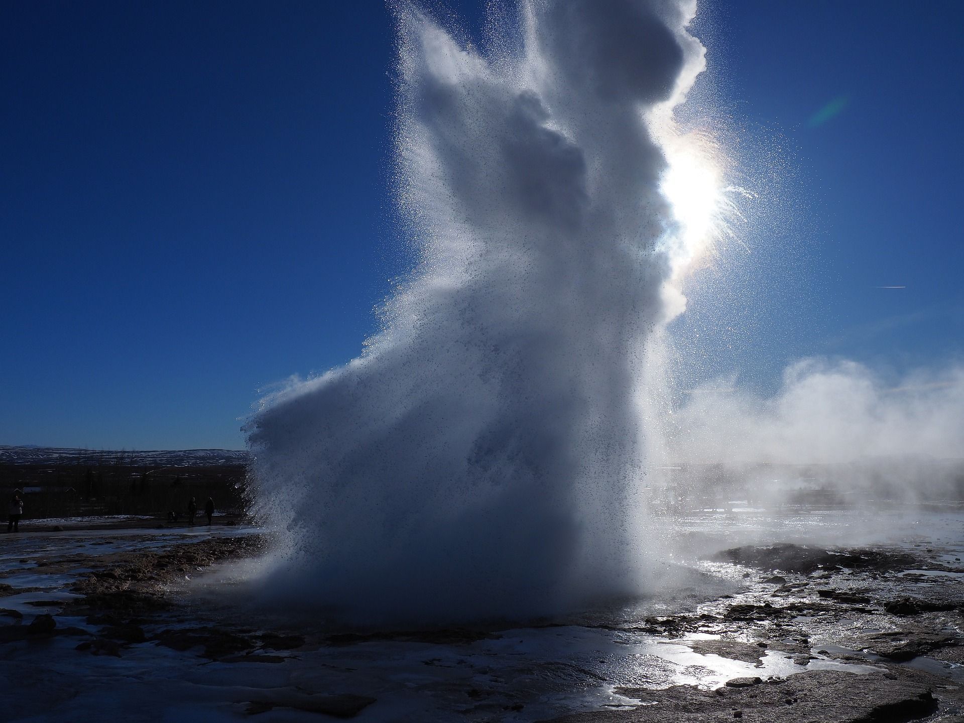 Ein großer Geysir bricht mit einem mächtigen Wasser- und Dampfstrahl aus, der von der Sonne vor einem klaren blauen Himmel hinterleuchtet wird.