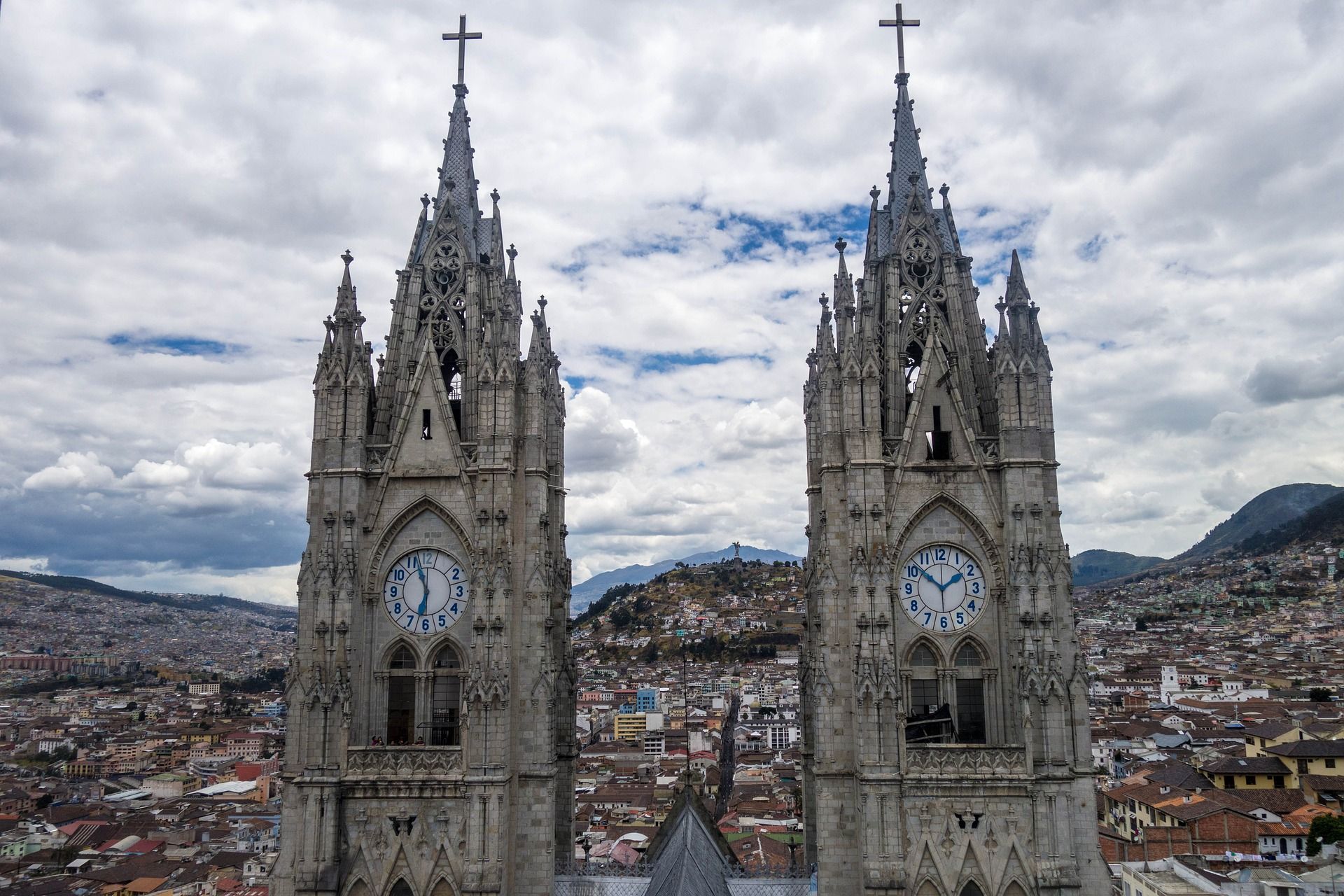 Dos torres de reloj de catedral de estilo gótico dominan una ciudad extensa con colinas al fondo bajo un cielo nublado.