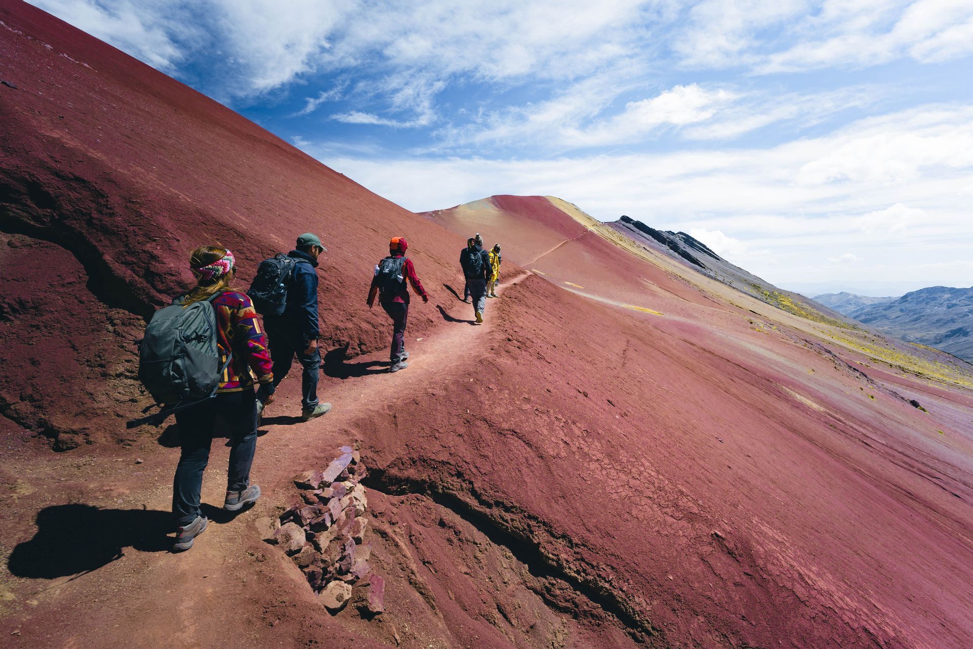 Un viaggio di gruppo WeRoad facendo trekking su uno stretto sentiero lungo il fianco di una vasta montagna rossa sotto un cielo blu parzialmente nuvoloso.