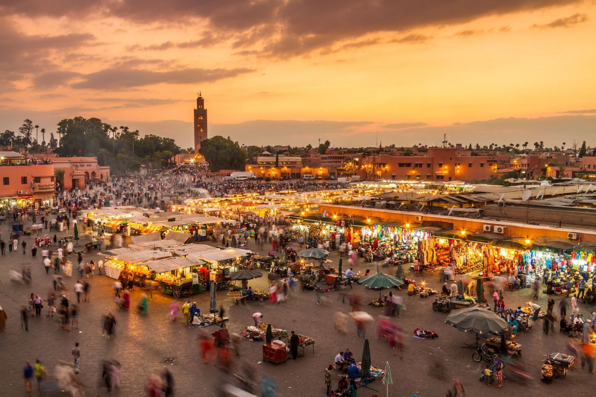 An aerial view of a bustling city market square filled with illuminated stalls and crowds of people at sunset.