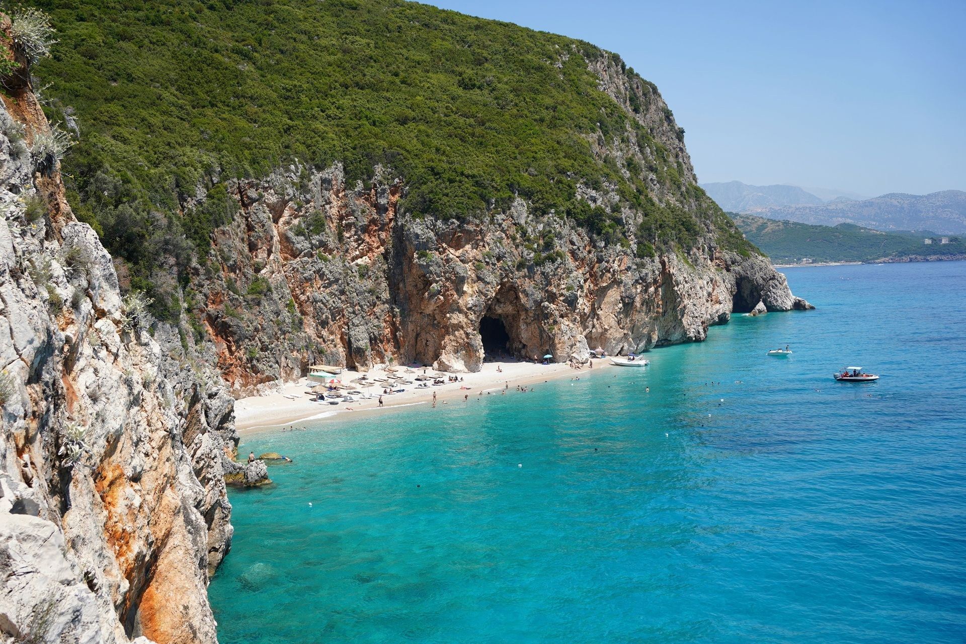 Una piccola spiaggia di sabbia con una grande grotta alla base di una scogliera verde e ripida lungo un'acqua turchese