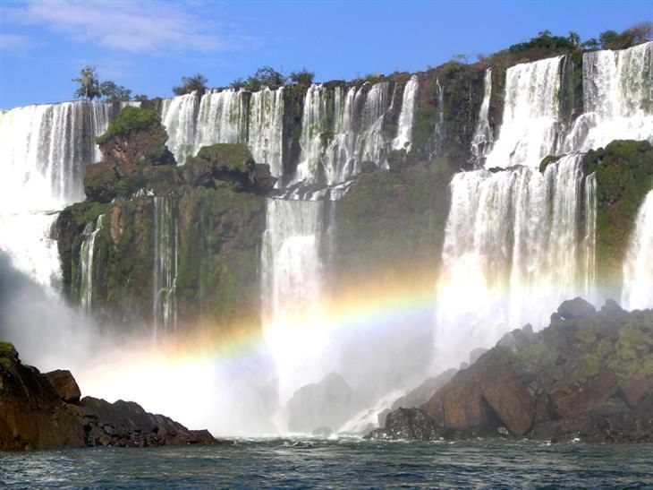 Una vasta cascata scende da una scogliera verde e rocciosa, creando un arcobaleno nella nebbia alla sua base.