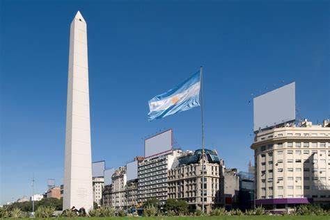 La bandiera argentina sventola accanto a un alto obelisco bianco in una piazza cittadina, con edifici storici sotto un cielo azzurro chiaro.