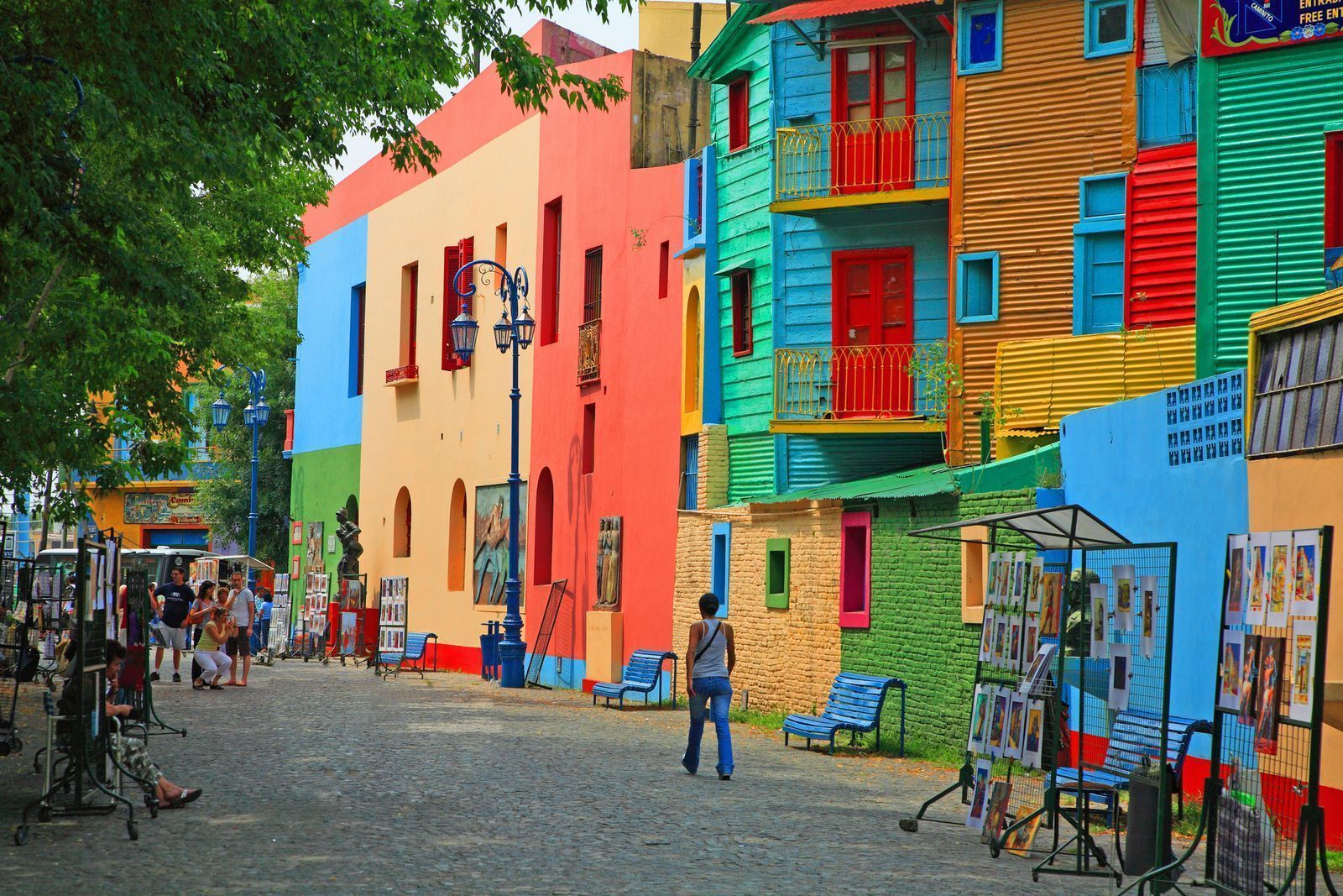 Una calle adoquinada, bañada por el sol, está bordeada de edificios vibrantes y multicolores mientras la gente camina y explora puestos de arte al aire libre.