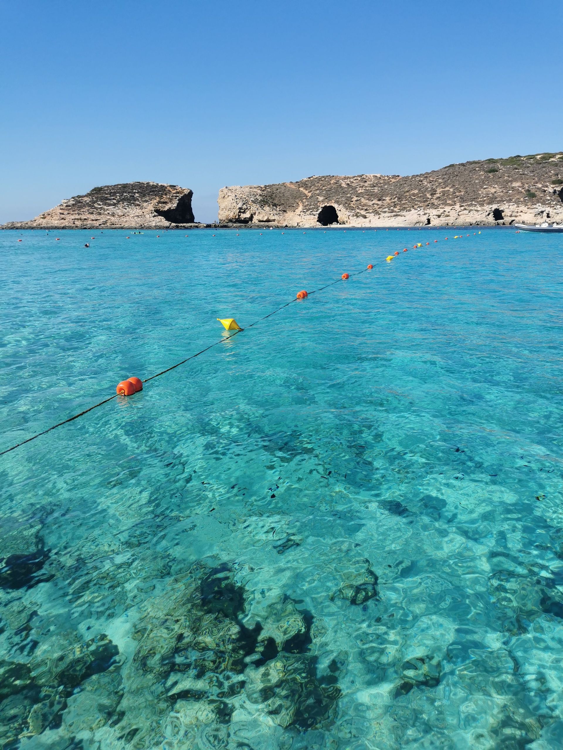 A line of floating buoys stretches across clear turquoise water toward rocky cliffs under a bright blue sky.