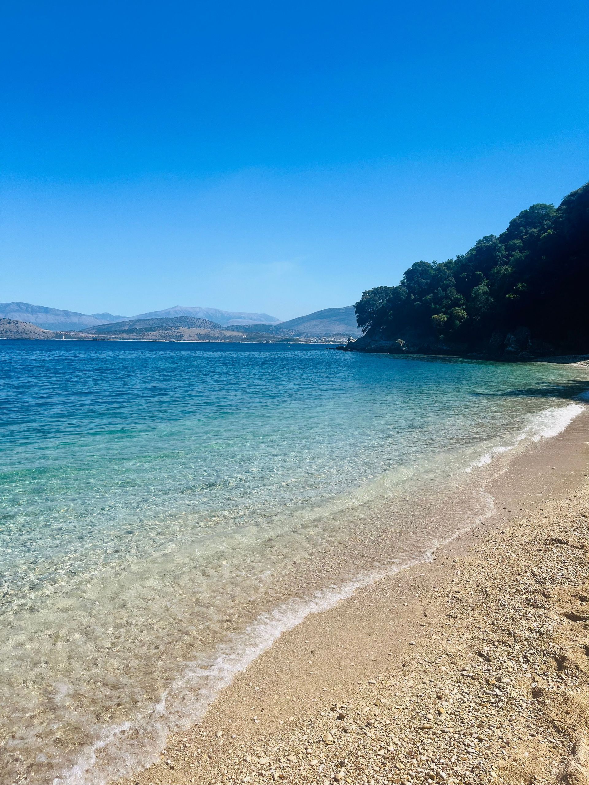 Clear turquoise water laps gently onto a pebble beach near forested hills under a bright blue sky.