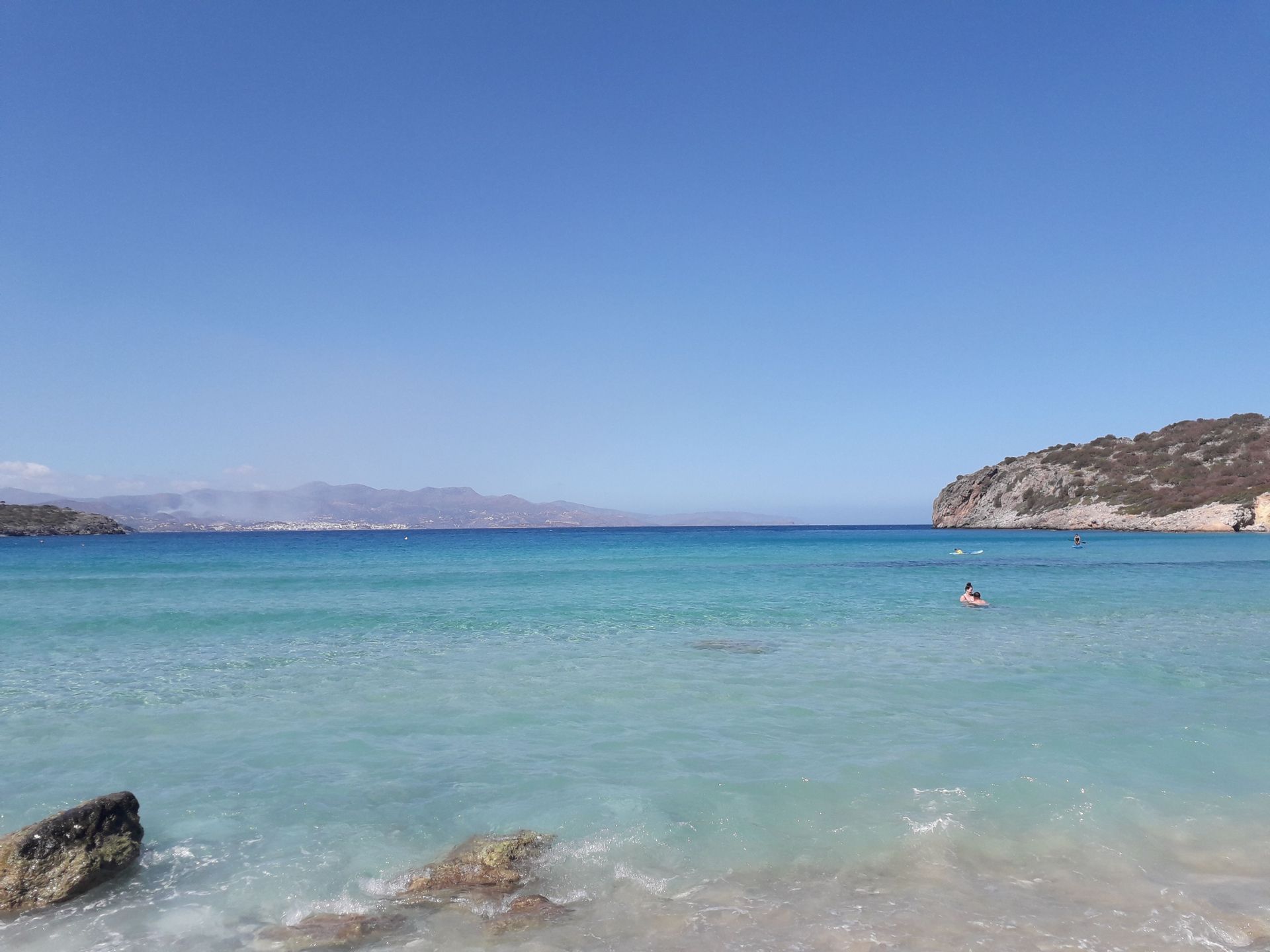 Clear turquoise sea waters in a coastal bay under a bright blue sky with hills in the background.