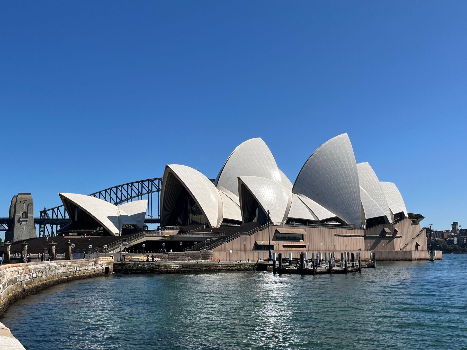 Un moderno edificio sul lungomare con tetti bianchi a forma di vela si erge accanto all'acqua blu con un grande ponte in acciaio ad arco sullo sfondo