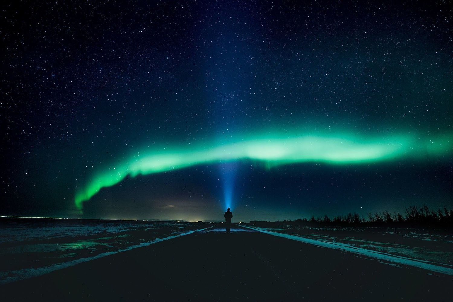 Una persona si trova su una strada di notte, illuminando con una torcia l'aurora boreale verde in un cielo stellato.