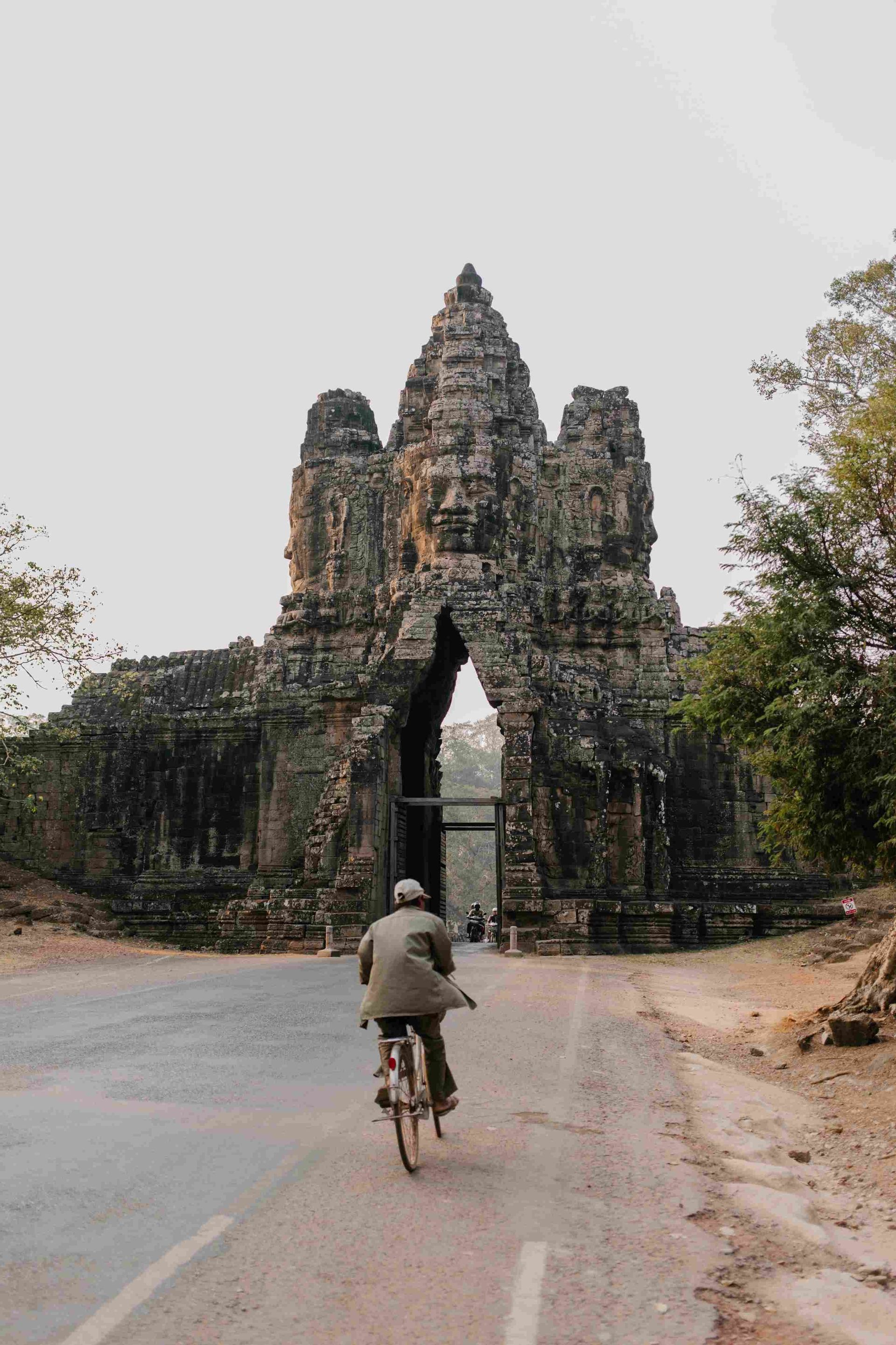 A person seen from behind rides a bicycle on a road towards a monumental ancient stone gate carved with large faces.