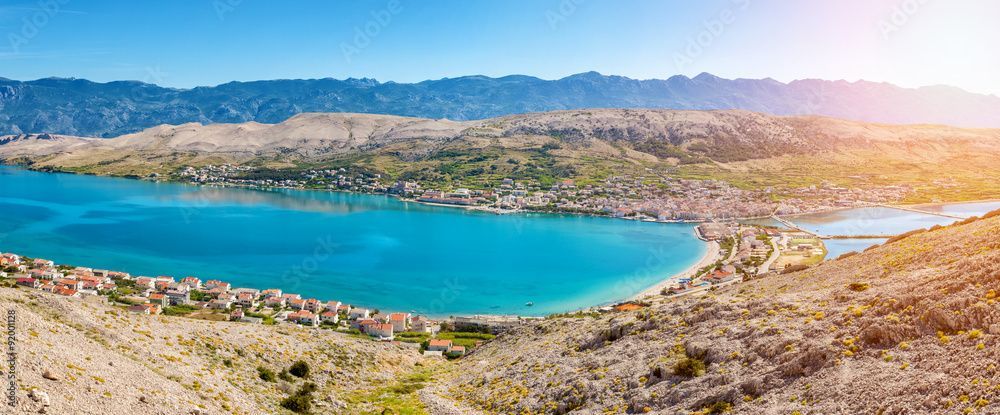 Panoramica di una cittadina costiera con case dai tetti rossi lungo una vivace baia blu, circondata da colline rocciose e montagne lontane.