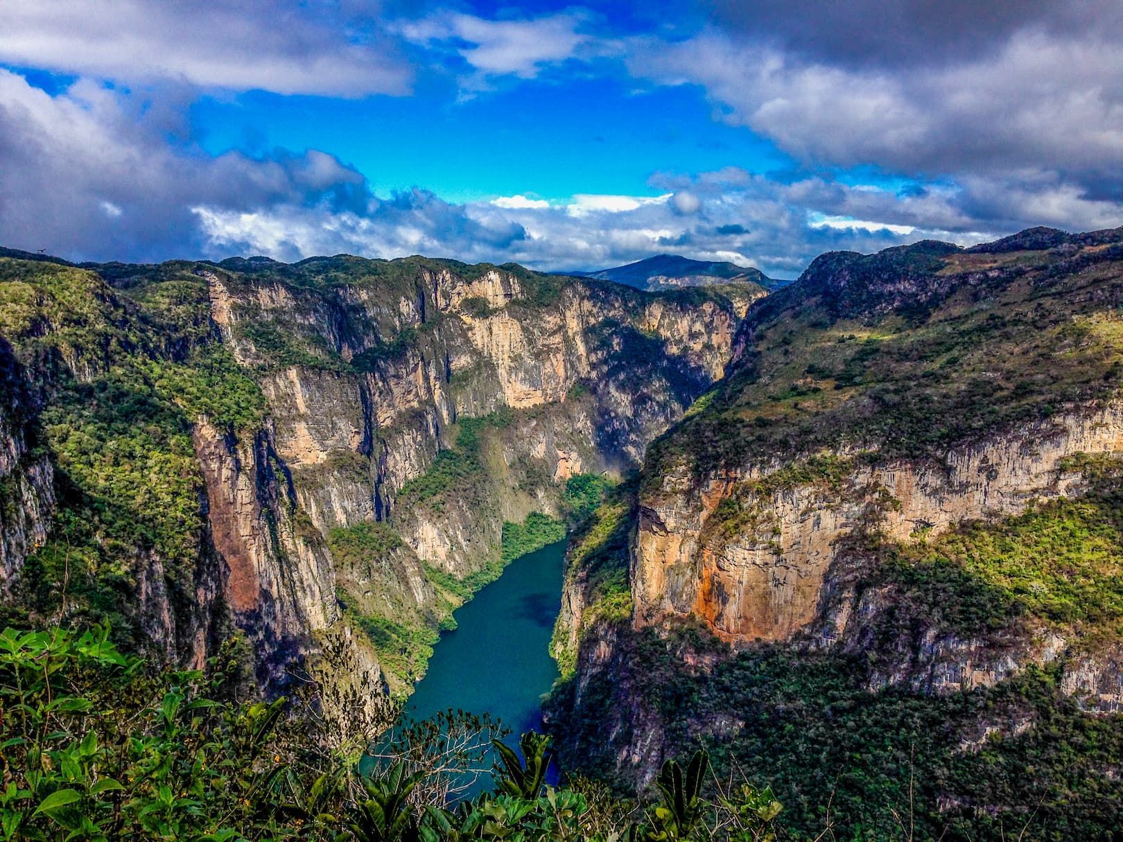 Ein türkiser Fluss fließt durch eine tiefe Schlucht mit steilen, vegetationsbedeckten Klippen unter einem blauen Himmel mit verstreuten Wolken.