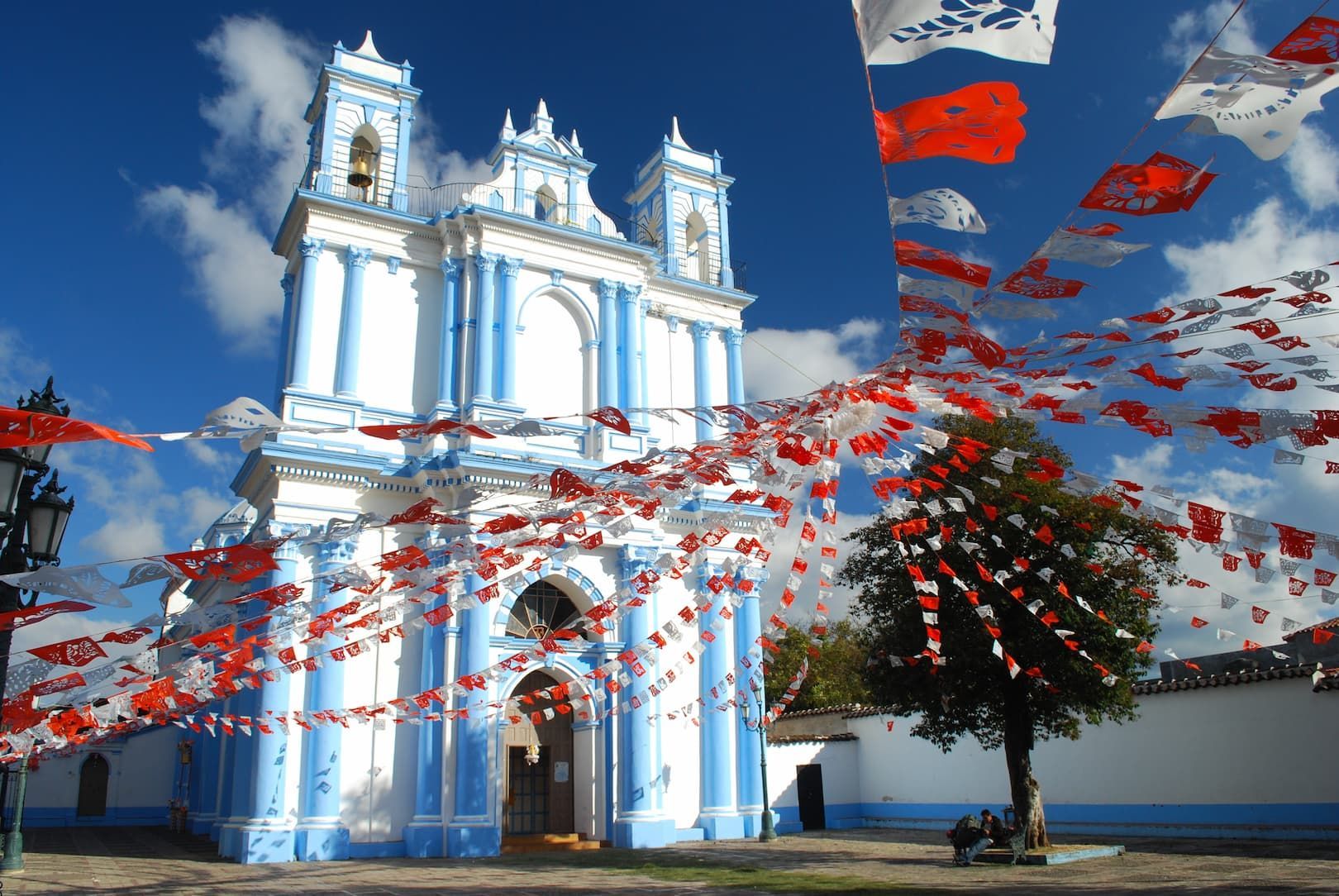 Una iglesia blanca y azul está decorada con guirnaldas de banderines de papel rojos y blancos en una plaza bajo un cielo azul claro.