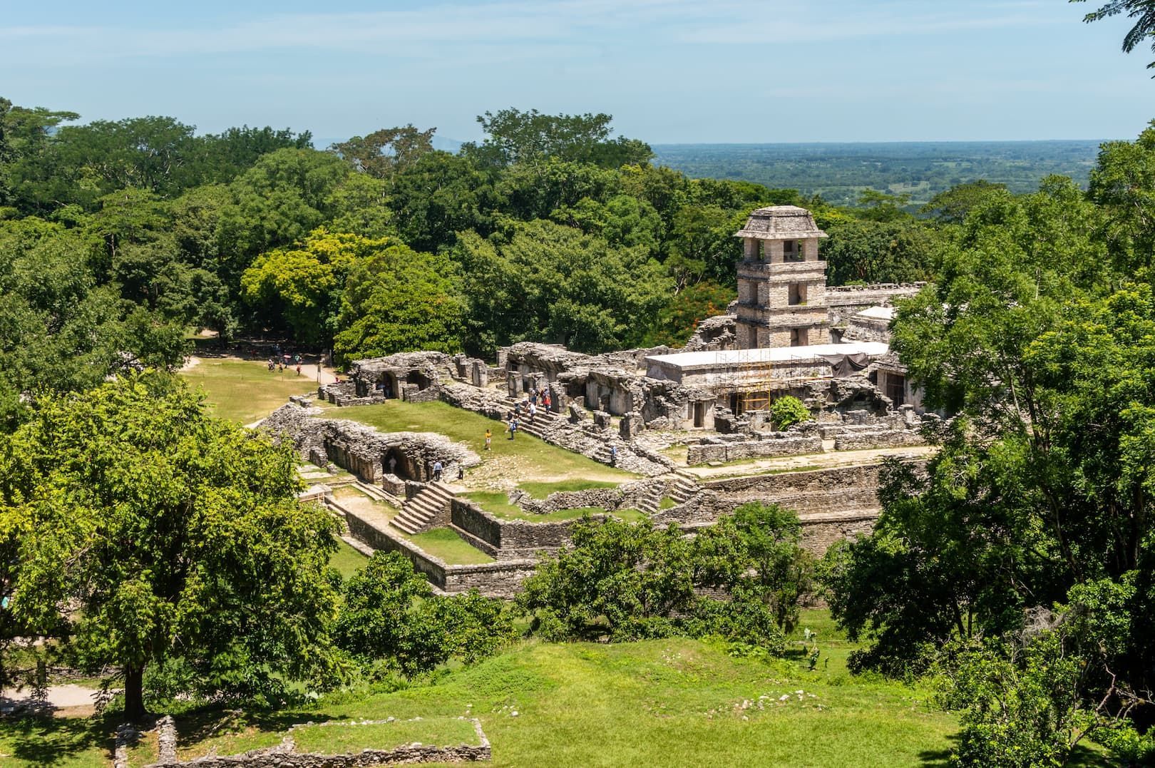 Vue grand-angle de ruines en pierre antiques, dont une tour proéminente, entourées d'une jungle verte dense sous un ciel clair.