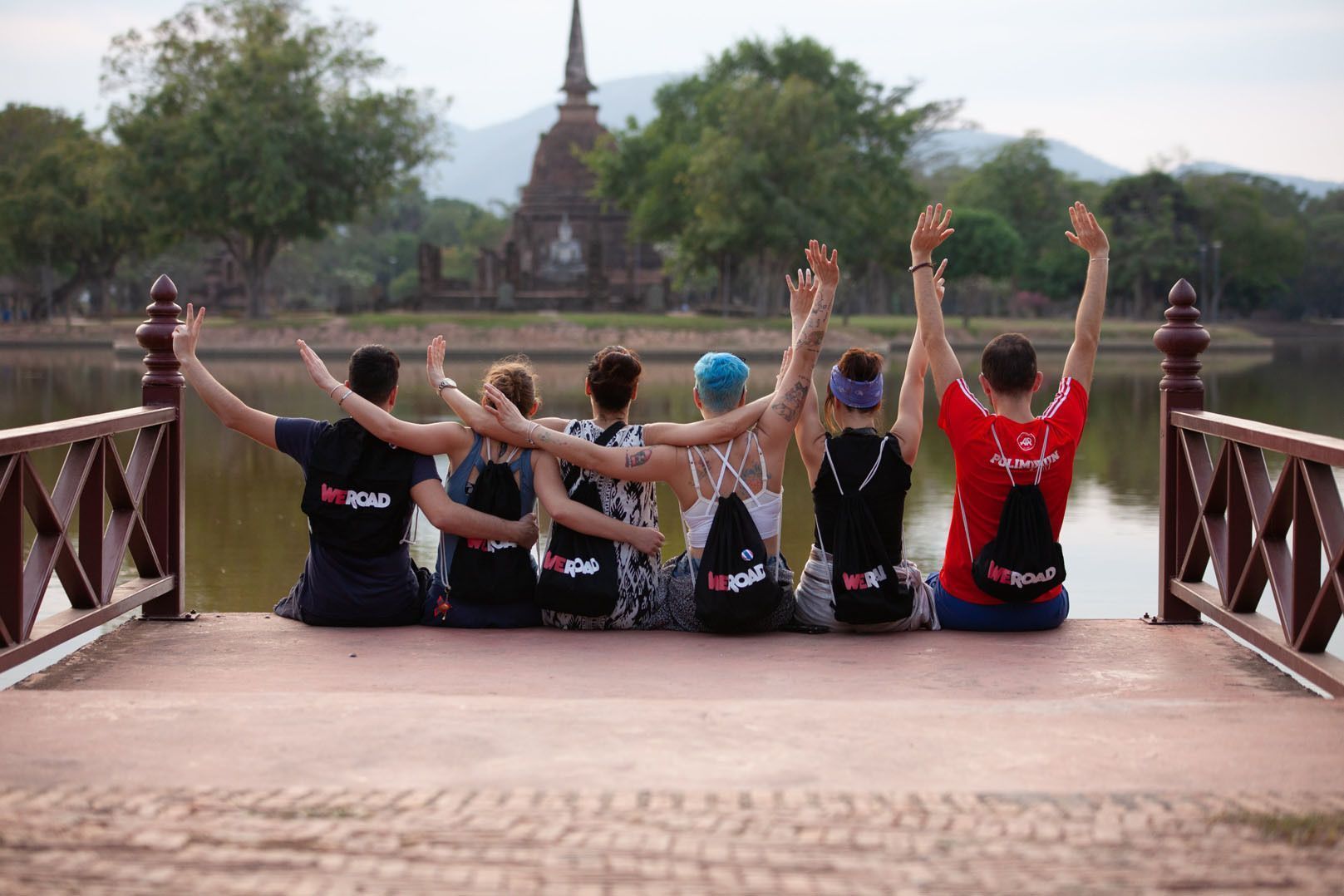 A WeRoad group trip sits on a pier with their arms raised, facing a historic temple across a body of water.