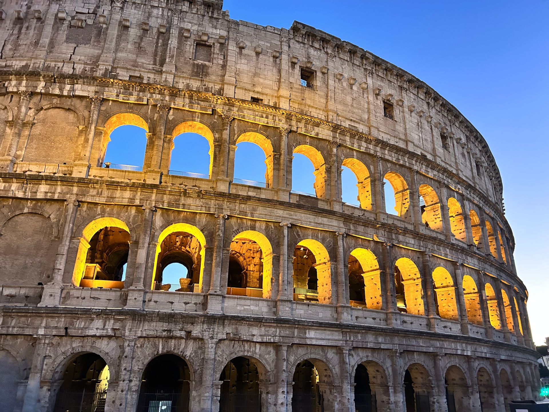 An ancient stone amphitheater with multiple tiers of arches illuminated by warm lights against a clear evening sky.