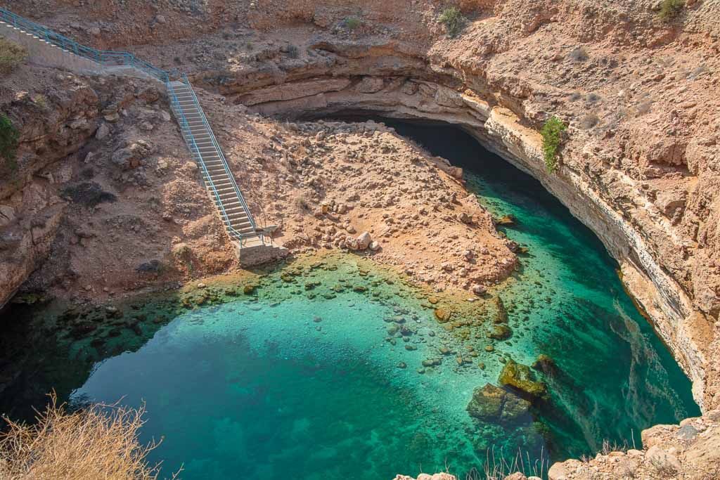 Una lunga scalinata scende in una conca rocciosa piena di acqua cristallina color turchese, incastonata tra scogliere aride.