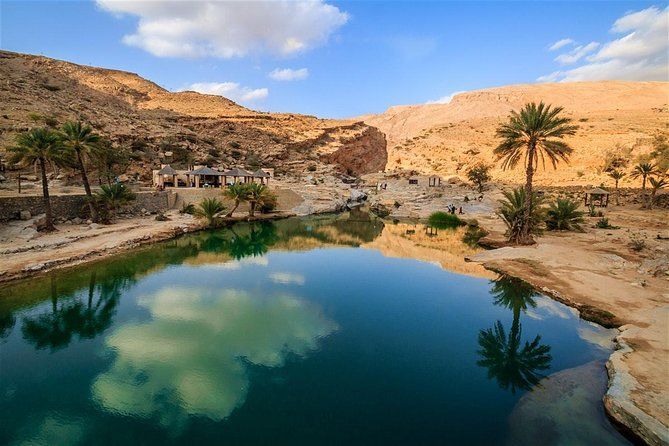 Un'oasi nel deserto con acqua calma e blu che riflette palme e un canyon roccioso sotto un cielo parzialmente nuvoloso.