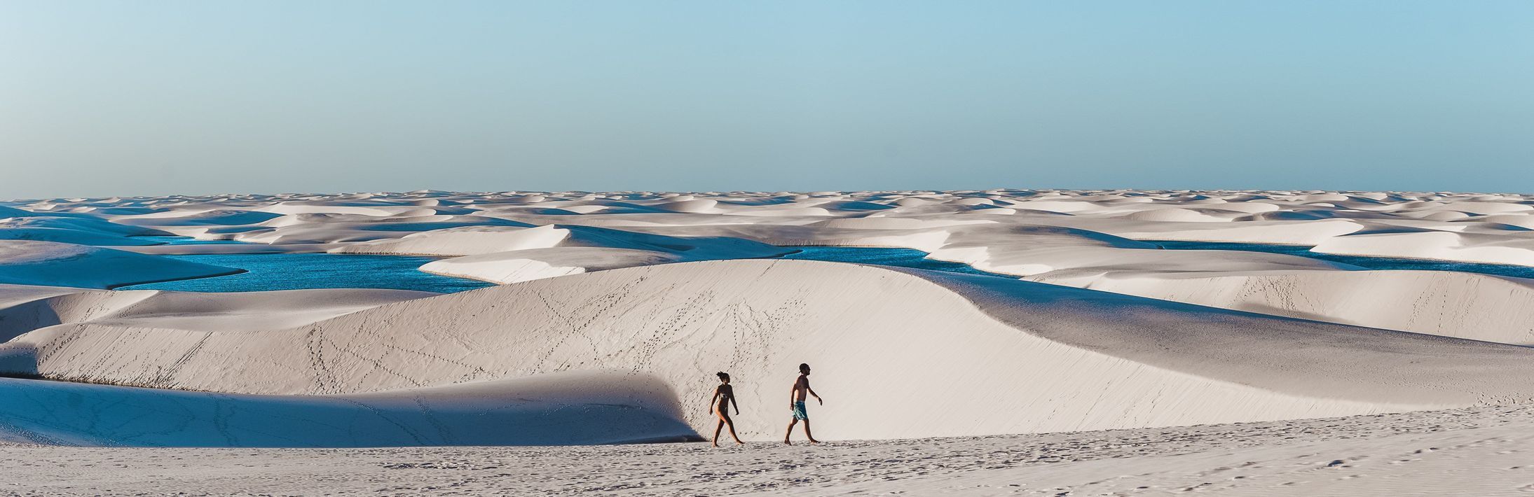 Zwei Personen gehen über weite, weiße Sanddünen, die mit blauen Lagunen gesprenkelt sind, unter einem klaren Himmel.