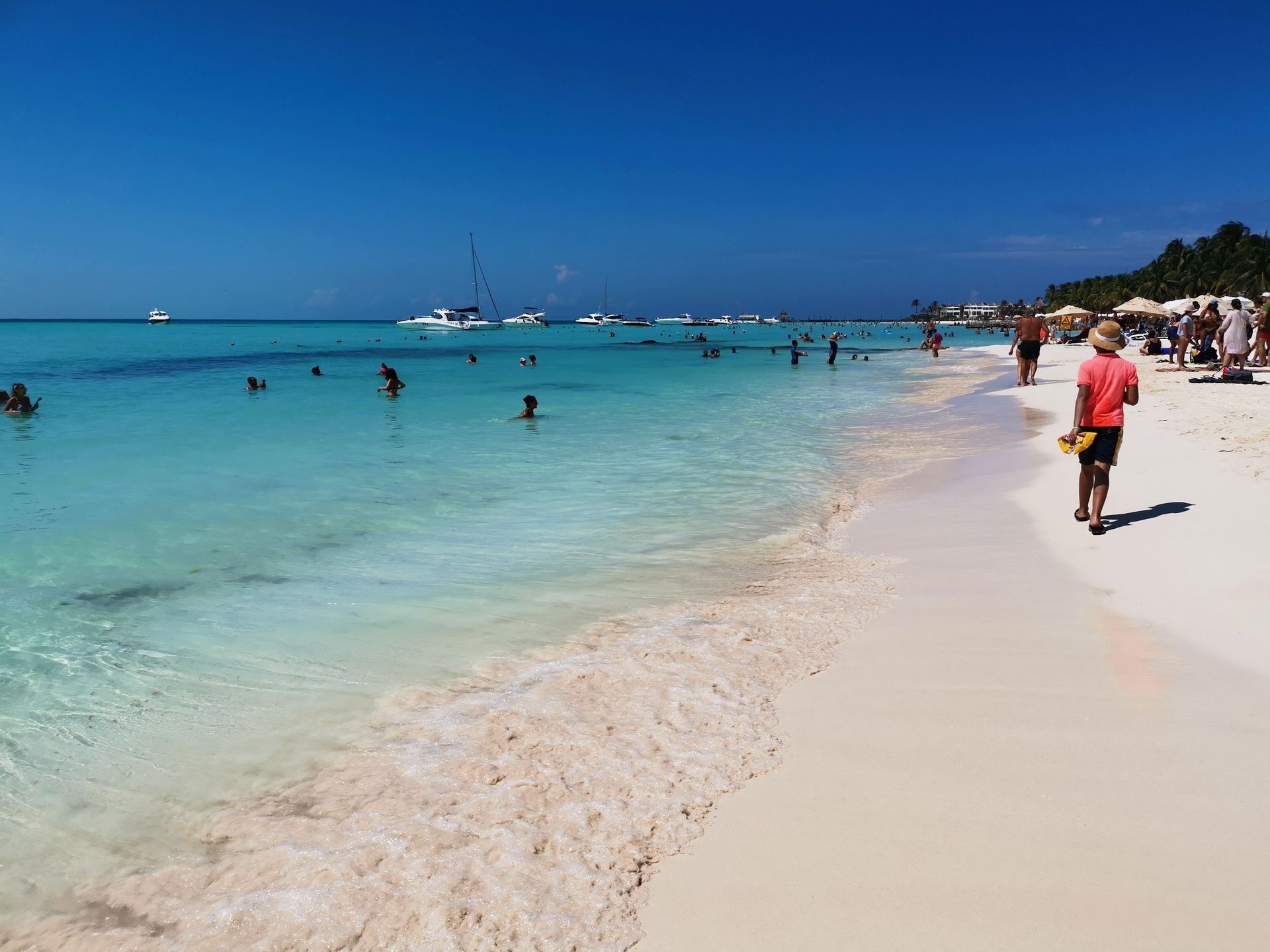 Many people enjoy a sunny day at a beach with white sand and clear turquoise water, with several boats on the horizon.