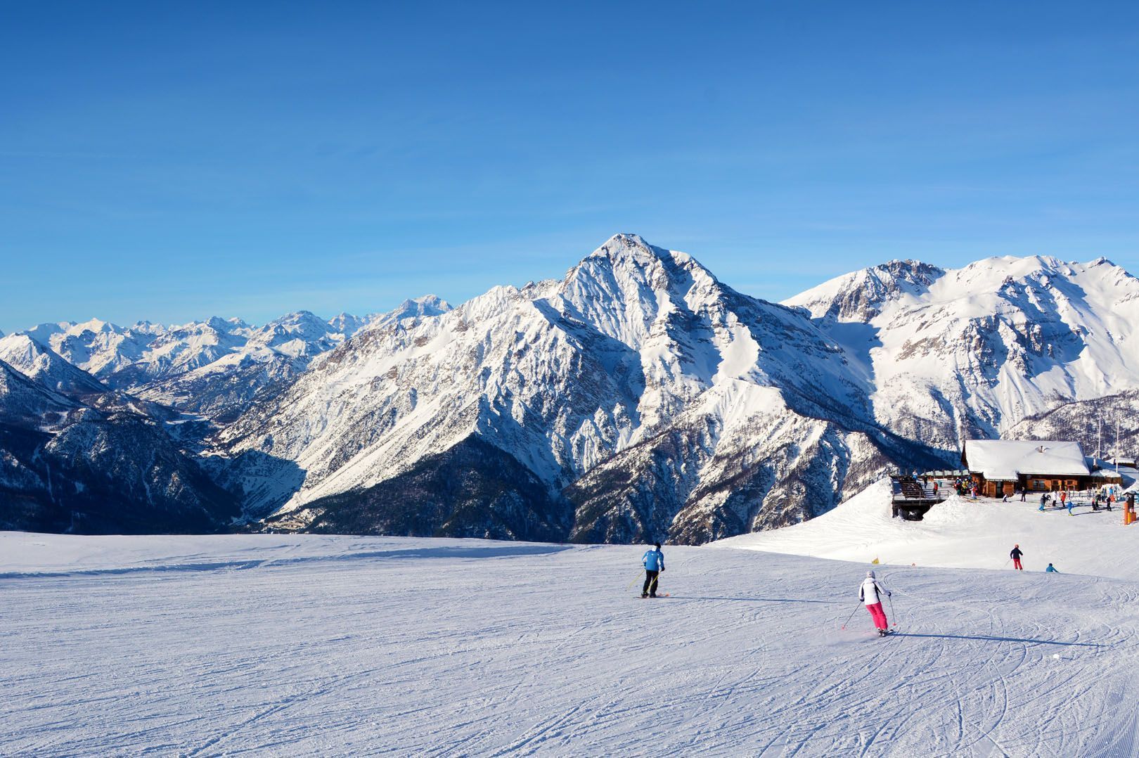 Personen beim Skifahren auf einer breiten, verschneiten Piste mit einer riesigen Bergkette und einer Skihütte im Hintergrund unter klarem blauem Himmel.