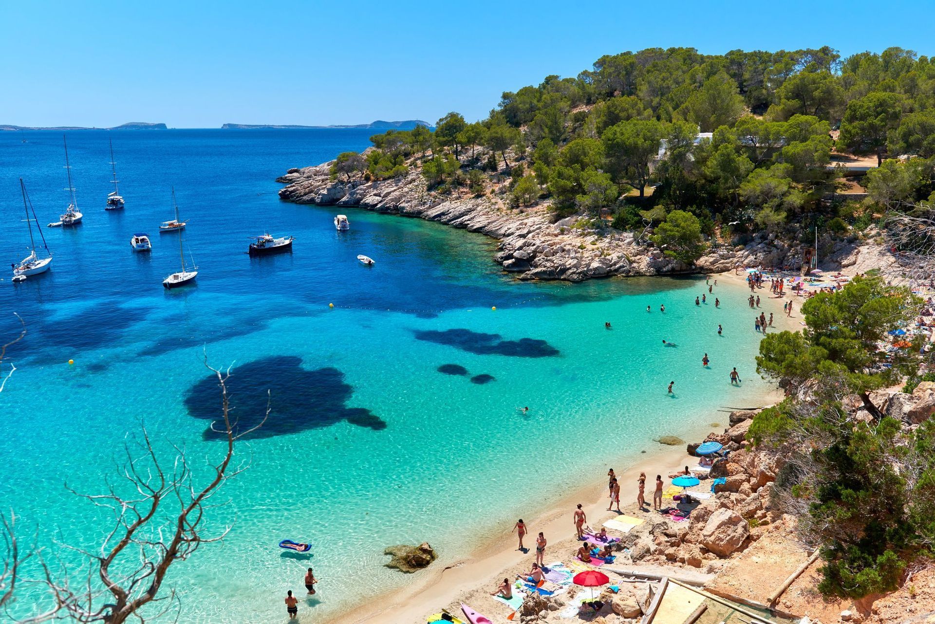 An aerial view of a busy beach cove with people swimming in turquoise water and boats anchored in the bay under a clear blue sky.