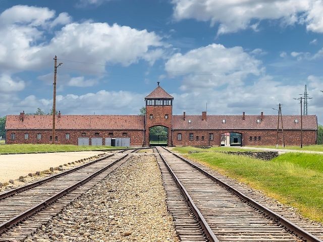 Dos conjuntos de vías de tren conducen hacia la entrada de un gran edificio de ladrillo con una torre de vigilancia central bajo un cielo azul con nubes.