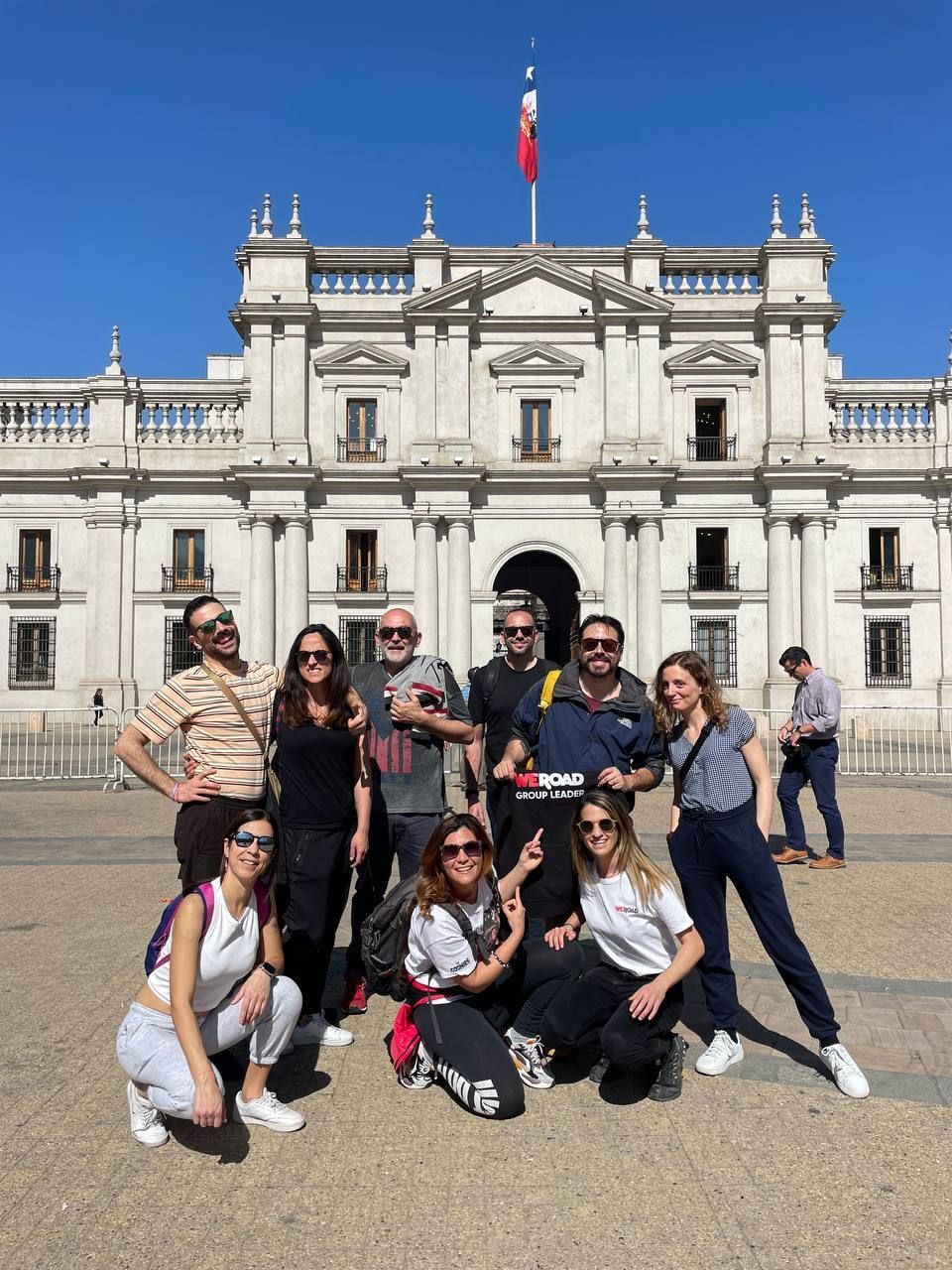 Un gruppo WeRoad in viaggio sorride per una foto in una piazza soleggiata, davanti a un imponente edificio classico con una bandiera.