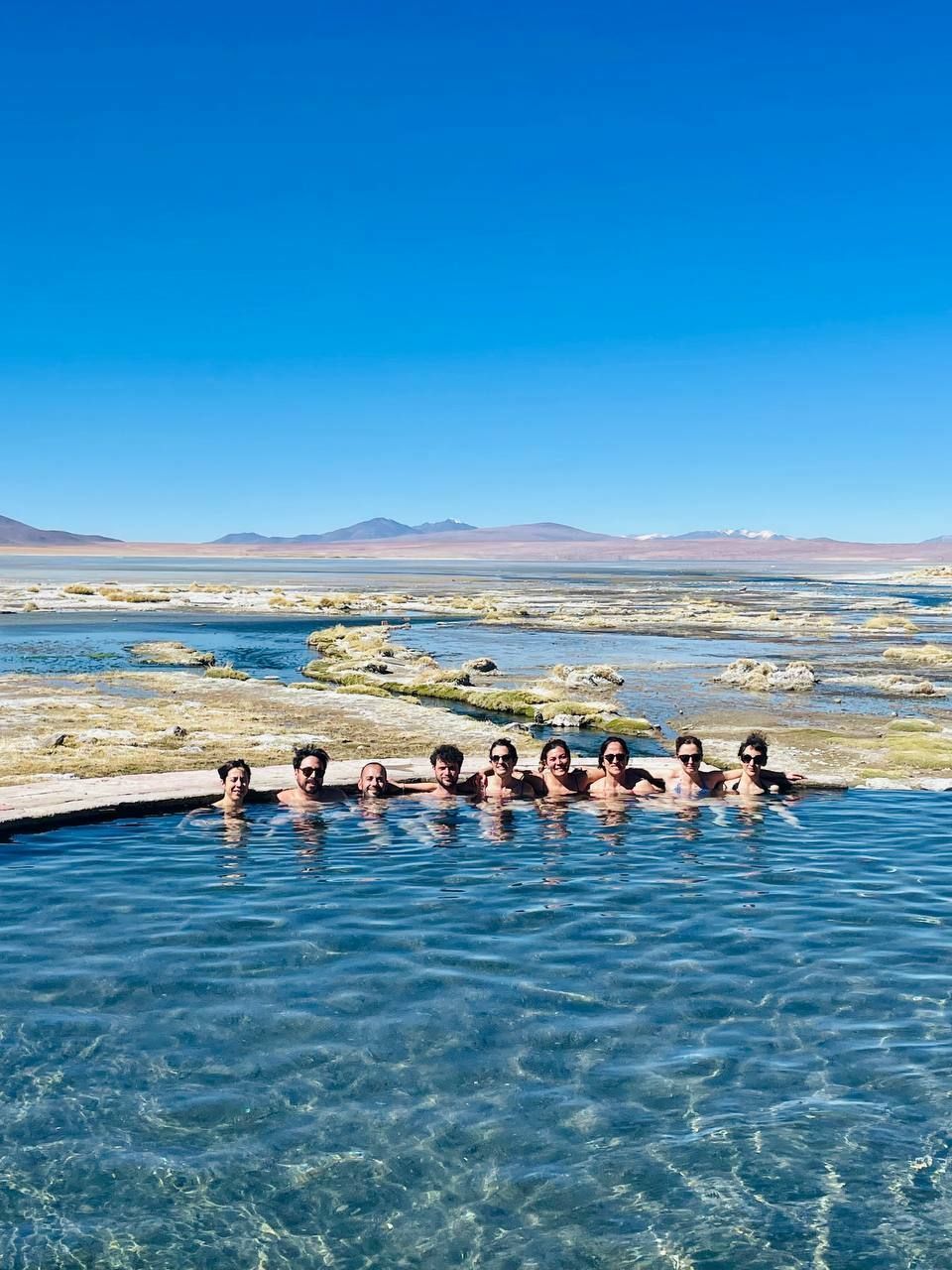 A WeRoad group trip posing for a photo while relaxing in a natural hot spring, with a vast, arid landscape in the background.