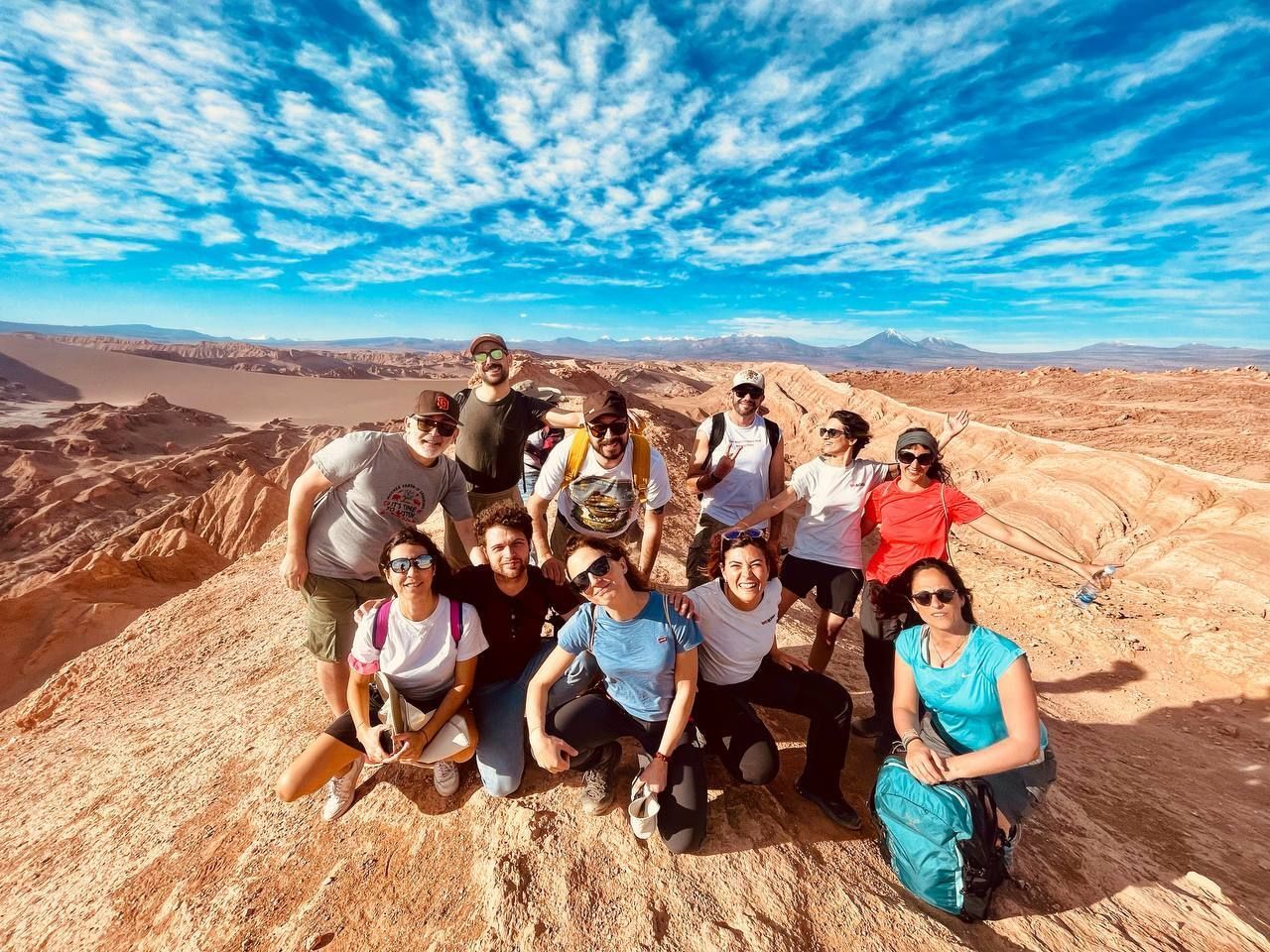 A WeRoad group trip poses for a photo on a rocky peak overlooking a desert landscape under a bright blue, cloudy sky.