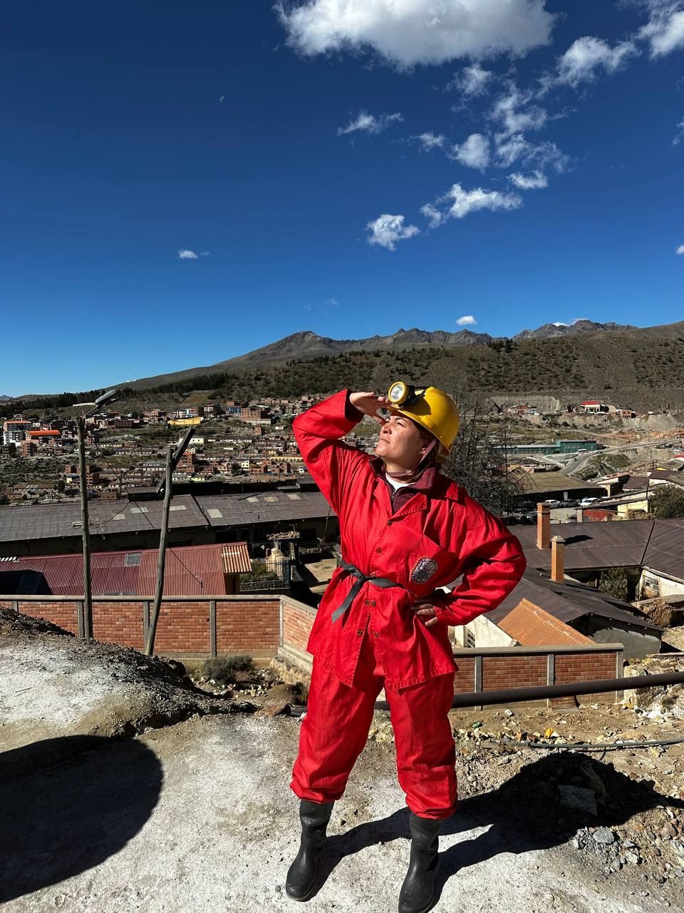 A woman wearing a red protective suit and a yellow hard hat with a headlamp stands on a hill overlooking a mining town.