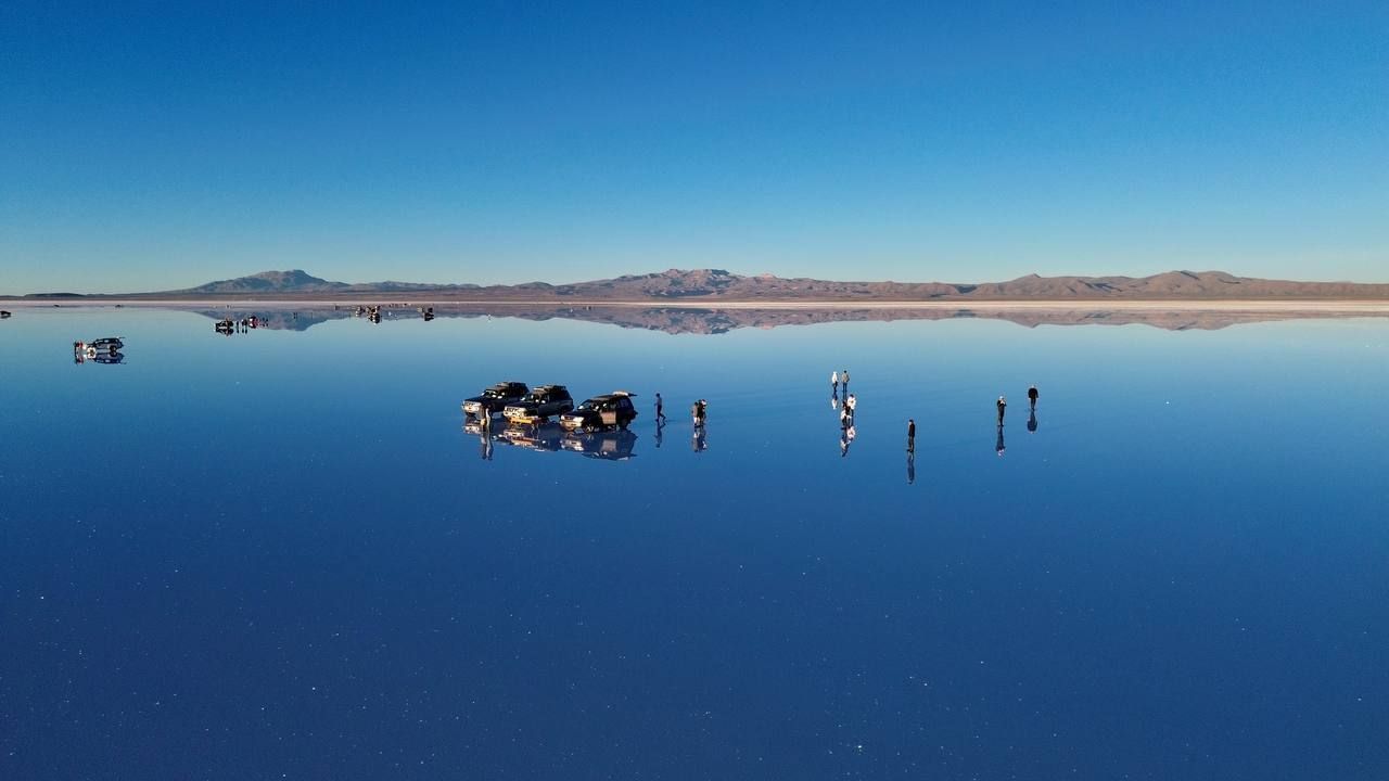 A WeRoad group trip stands on a vast, reflective salt flat with 4x4 vehicles, creating a perfect mirror of the blue sky and mountains.