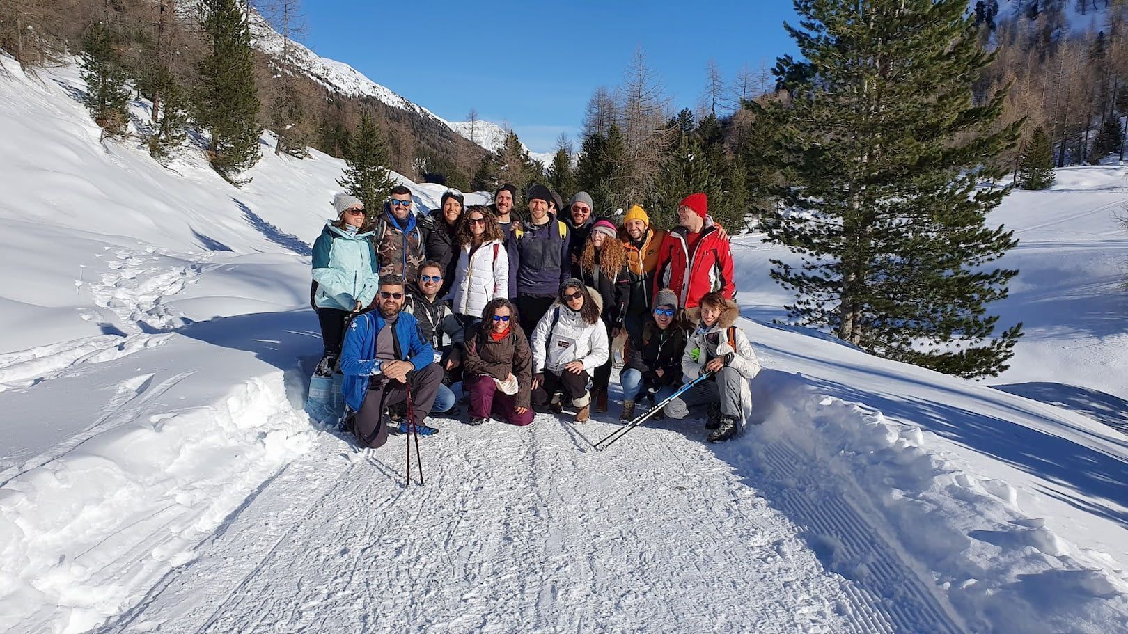 Un gruppo WeRoad in viaggio posa per una foto su un sentiero in una valle di montagna innevata, circondata da pini, sotto un cielo azzurro e limpido.
