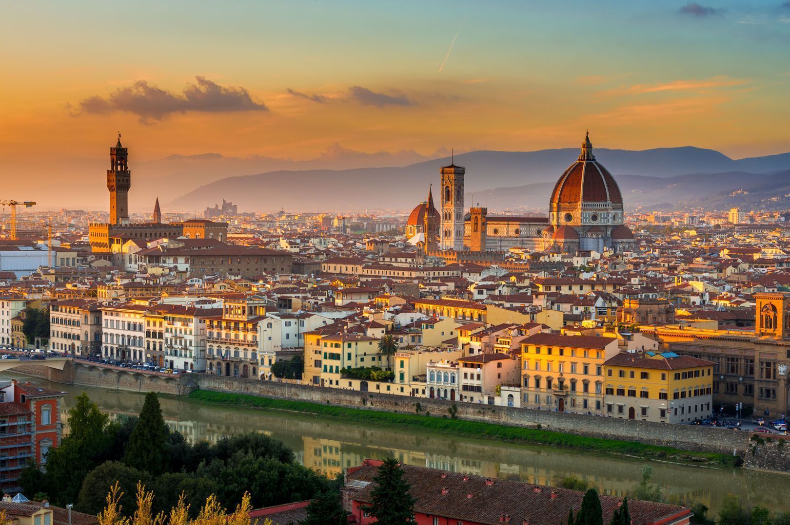 A panoramic view of a historic European city at sunset, featuring a prominent domed cathedral and tower along a river.